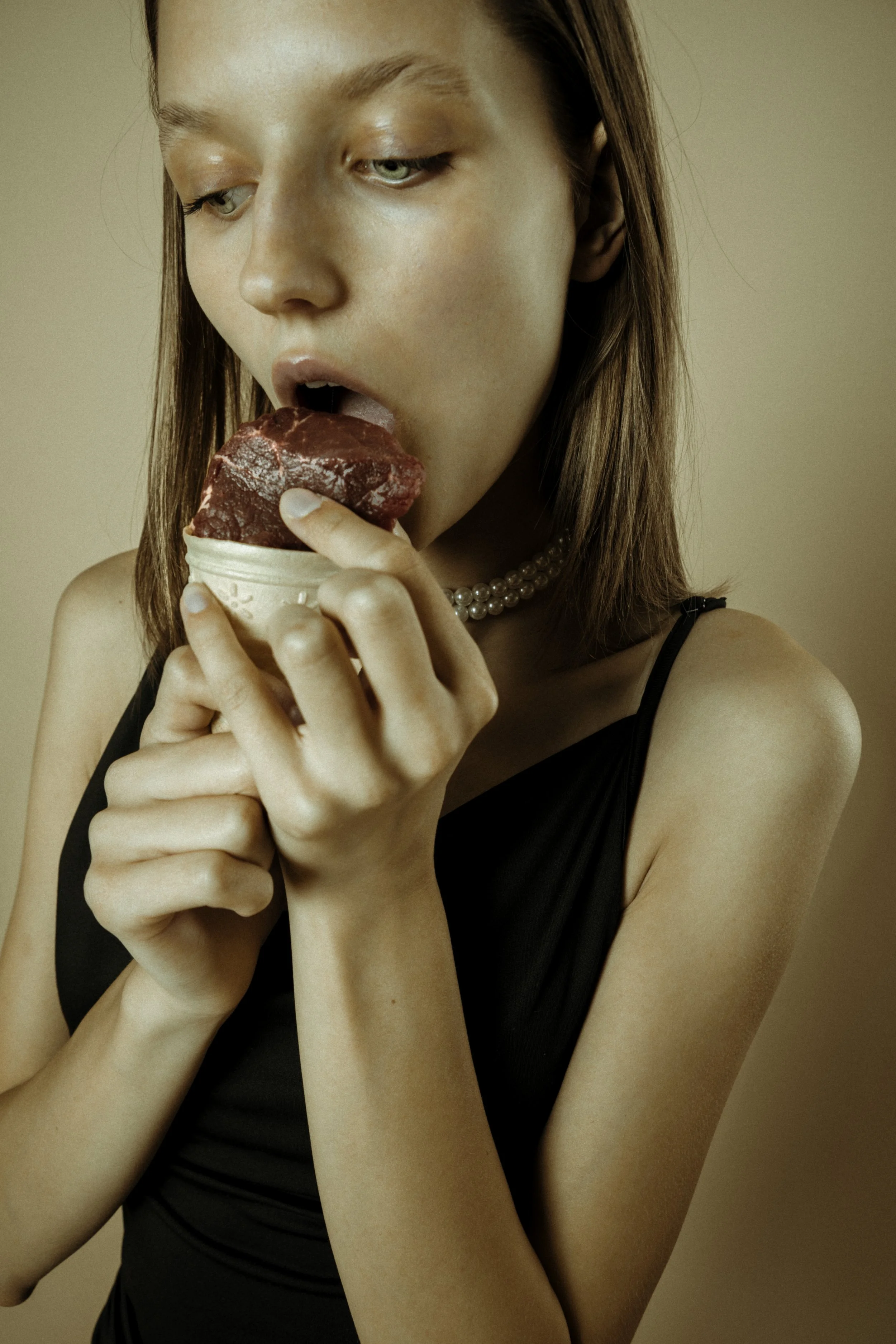 Woman in a black dress with pearl necklace licking a scoop of red ice cream in a cone.