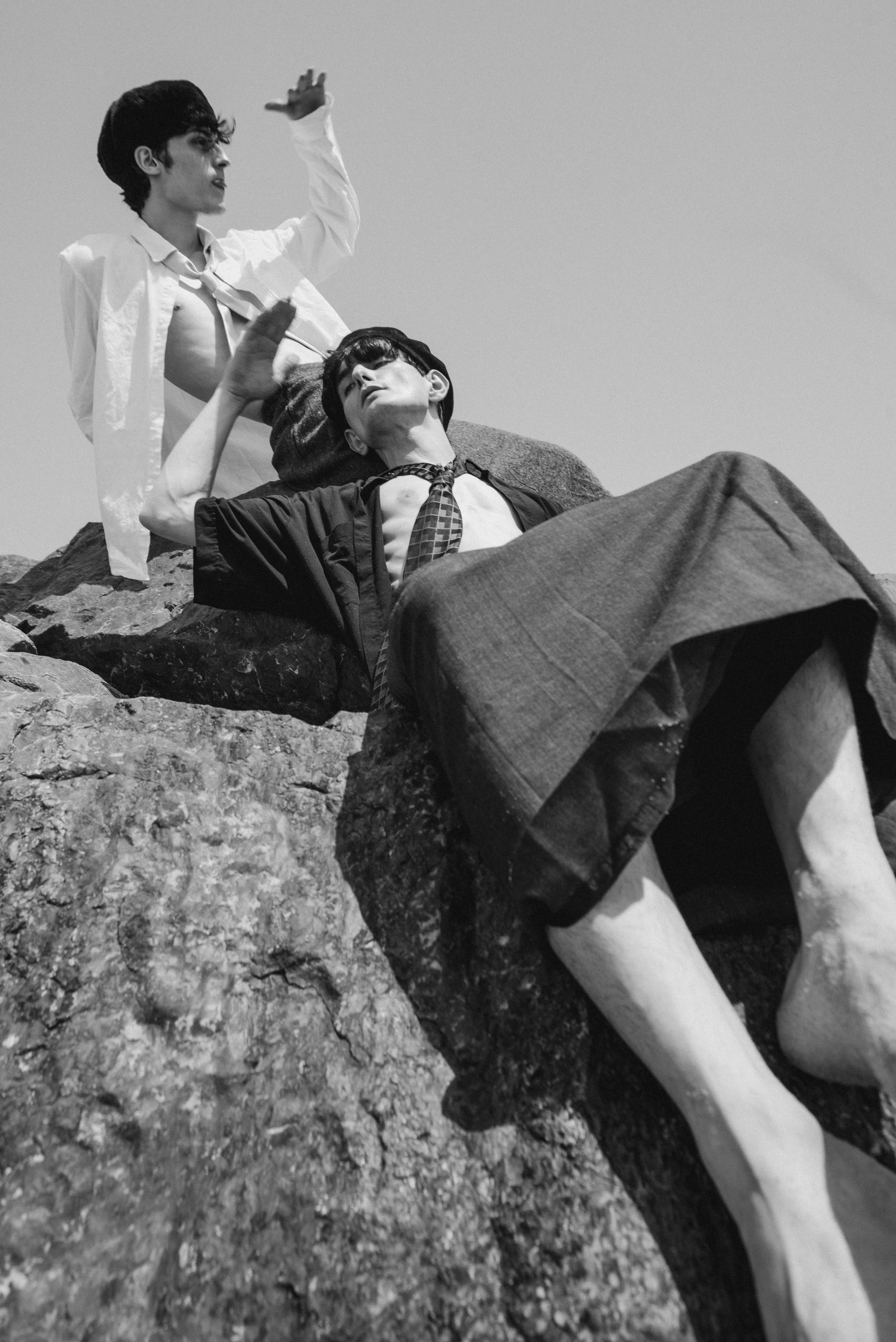 Two young men with dark hair, dressed in vintage clothing, are lying on rocks outdoors, one sitting upright and the other reclining, with another person nearby, all against a clear sky.