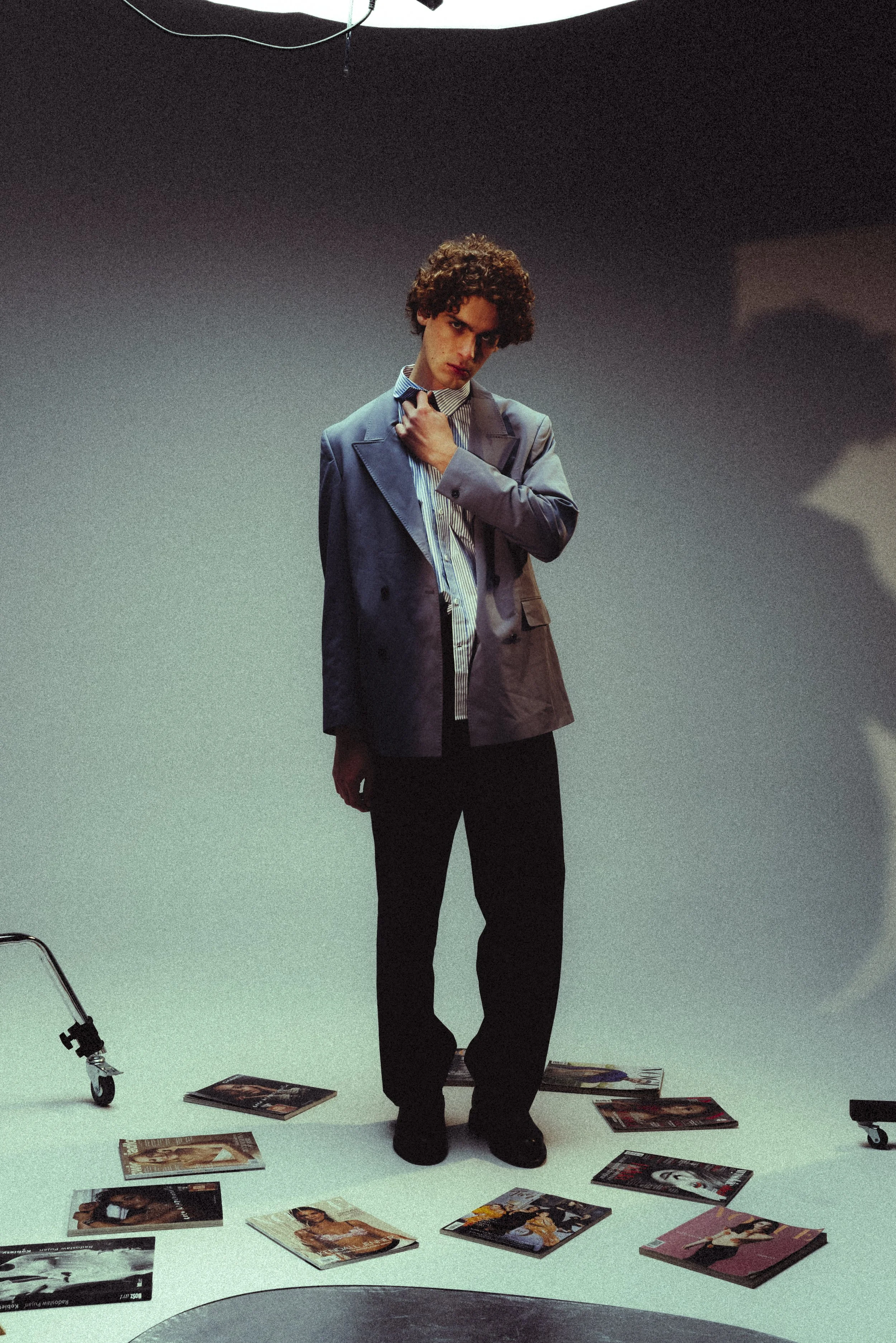 Young man with curly hair in a gray suit, standing in a photography studio with magazines scattered on the floor and a shopping cart in the background.