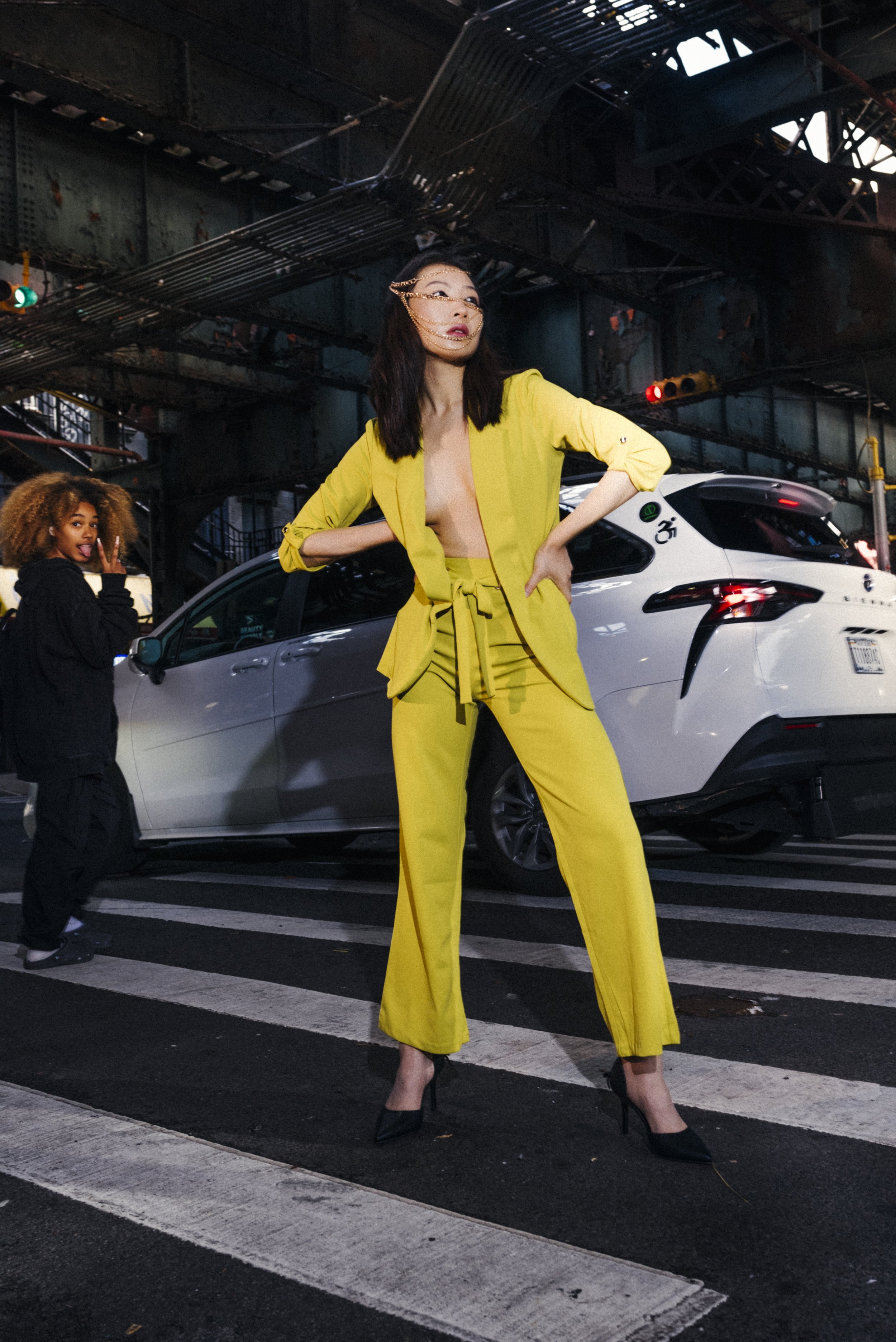 A woman in a yellow suit with high heels stands confidently on a crosswalk under a bridge in an urban setting, with a white car behind her and a young girl making a peace sign in the background.