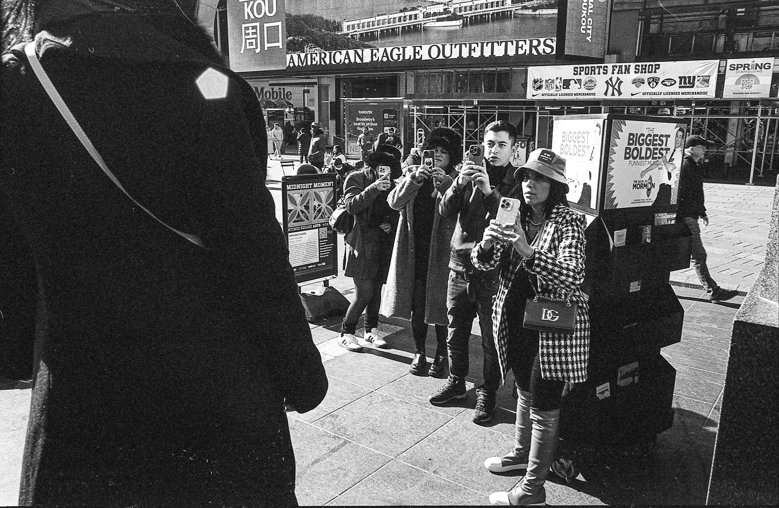A group of five people standing in a line on the sidewalk, taking photos or videos with their smartphones. They are in front of a building with signs advertising merchandise and a sports fan shop. There are other pedestrians walking by in the backgro