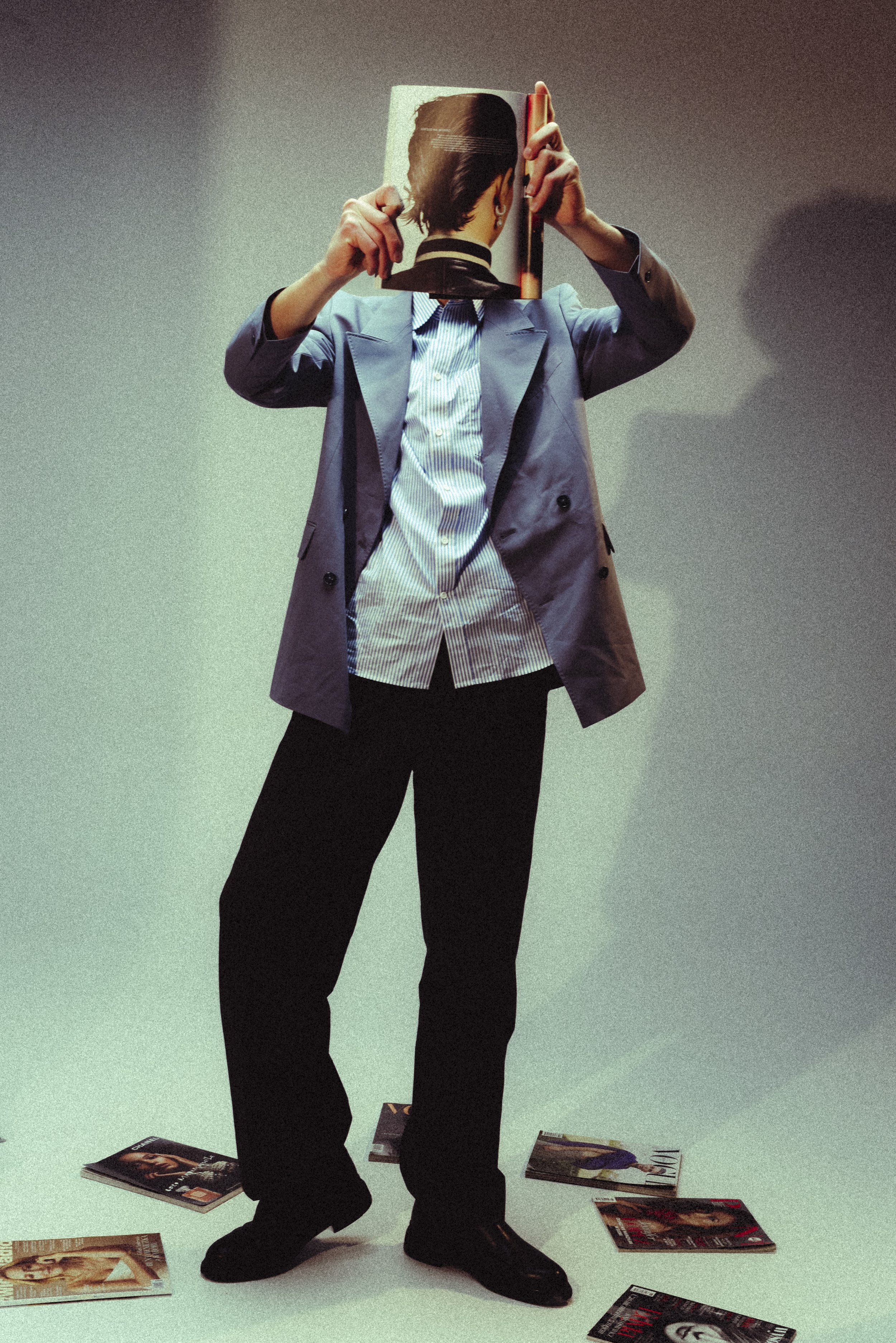 Person in a blue blazer and black pants holding a magazine in front of their face, with magazine pages scattered on the floor.