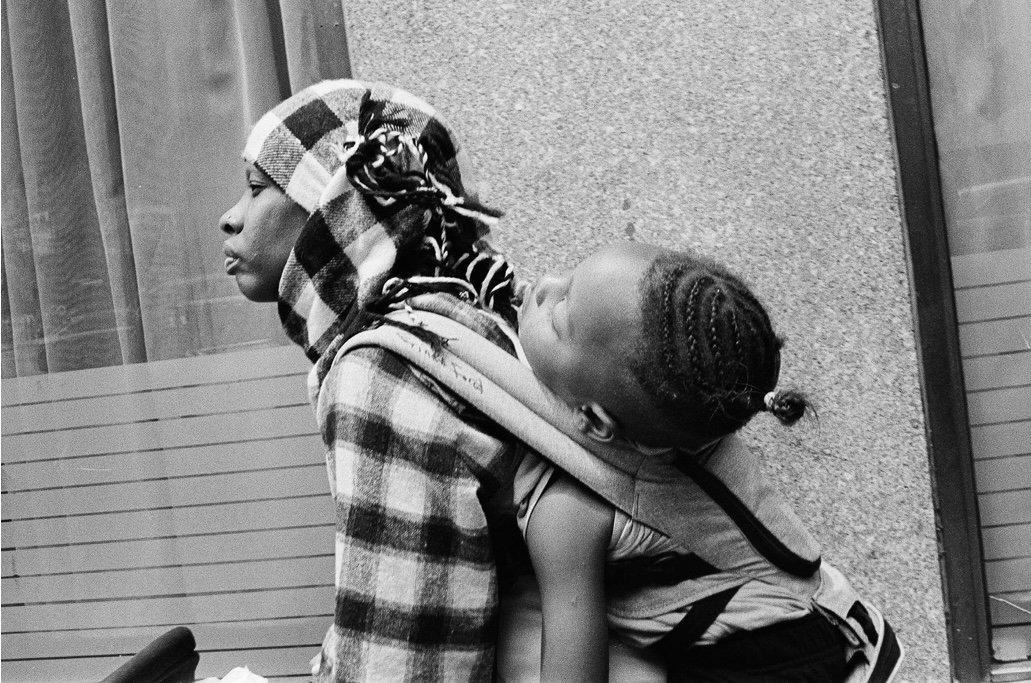 A woman carrying a young girl on her back, walking in front of a brick wall and glass windows.