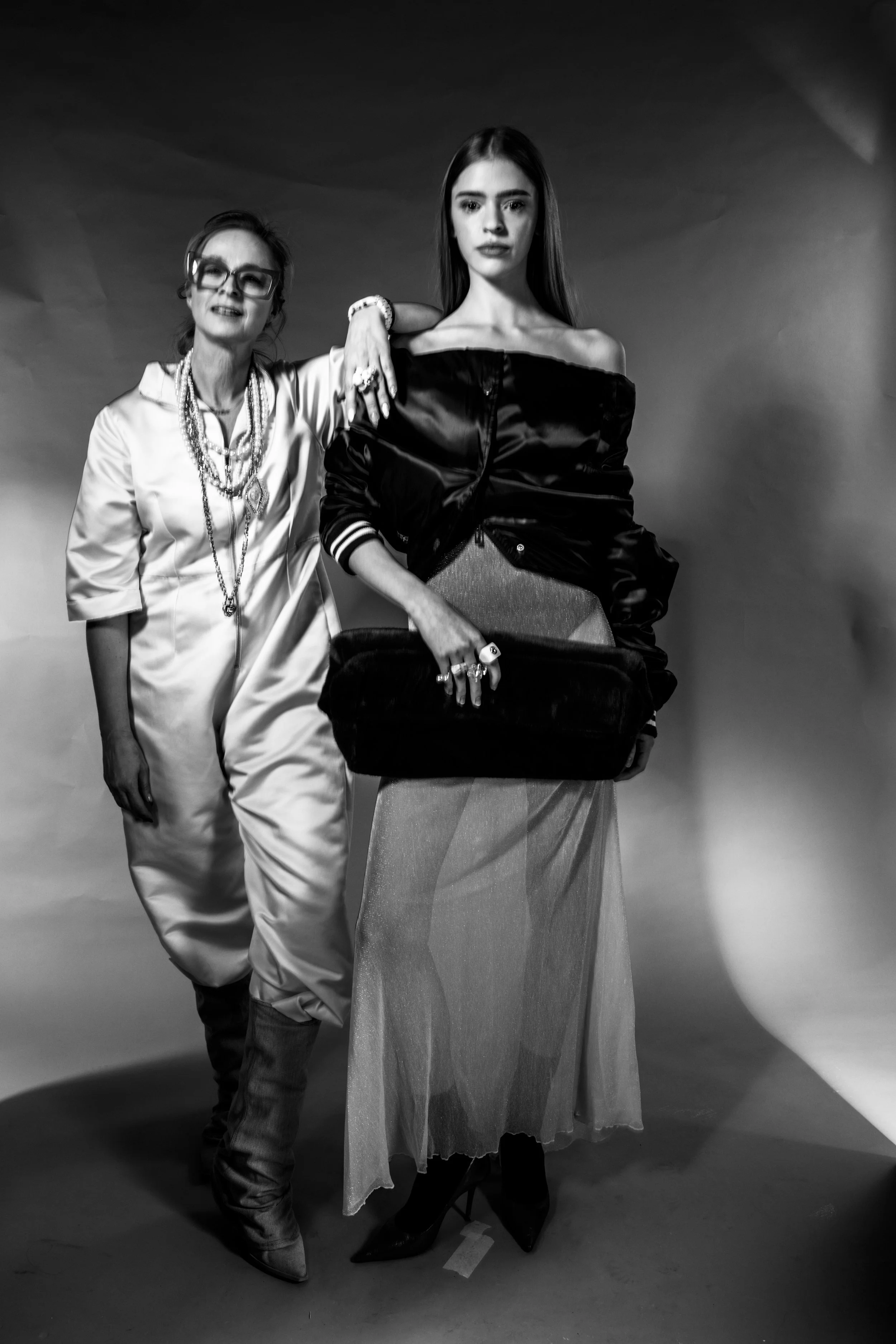 Two women posing for a black and white photo, one dressed in fashion-forward clothing with jewelry, holding a purse, and the other in a satin outfit with boots, against a plain backdrop.