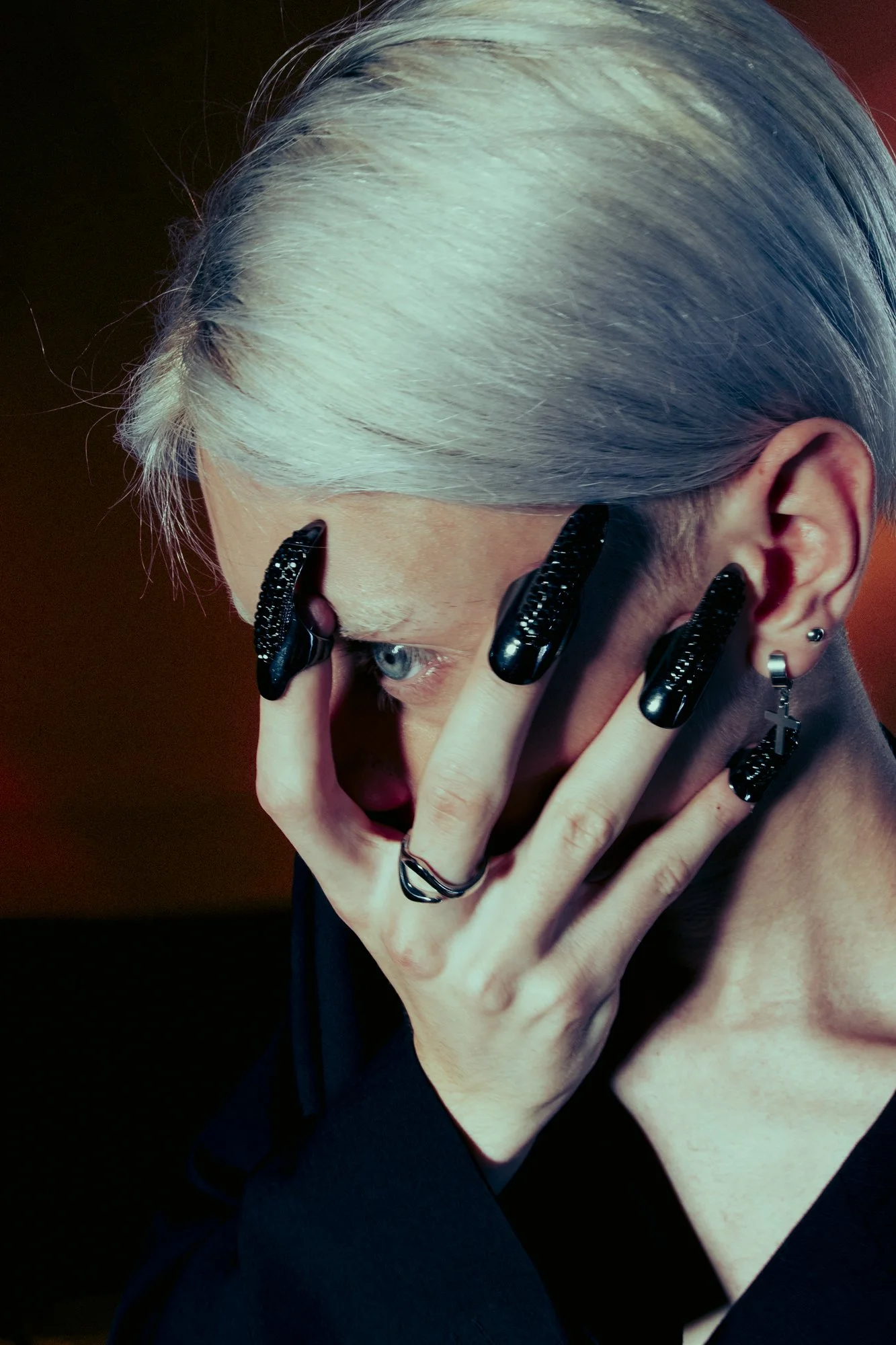 Close-up of a person with platinum blonde hair and blue eyes covering part of their face with their hand, which has black nail polish and jewelry, including earrings and rings.