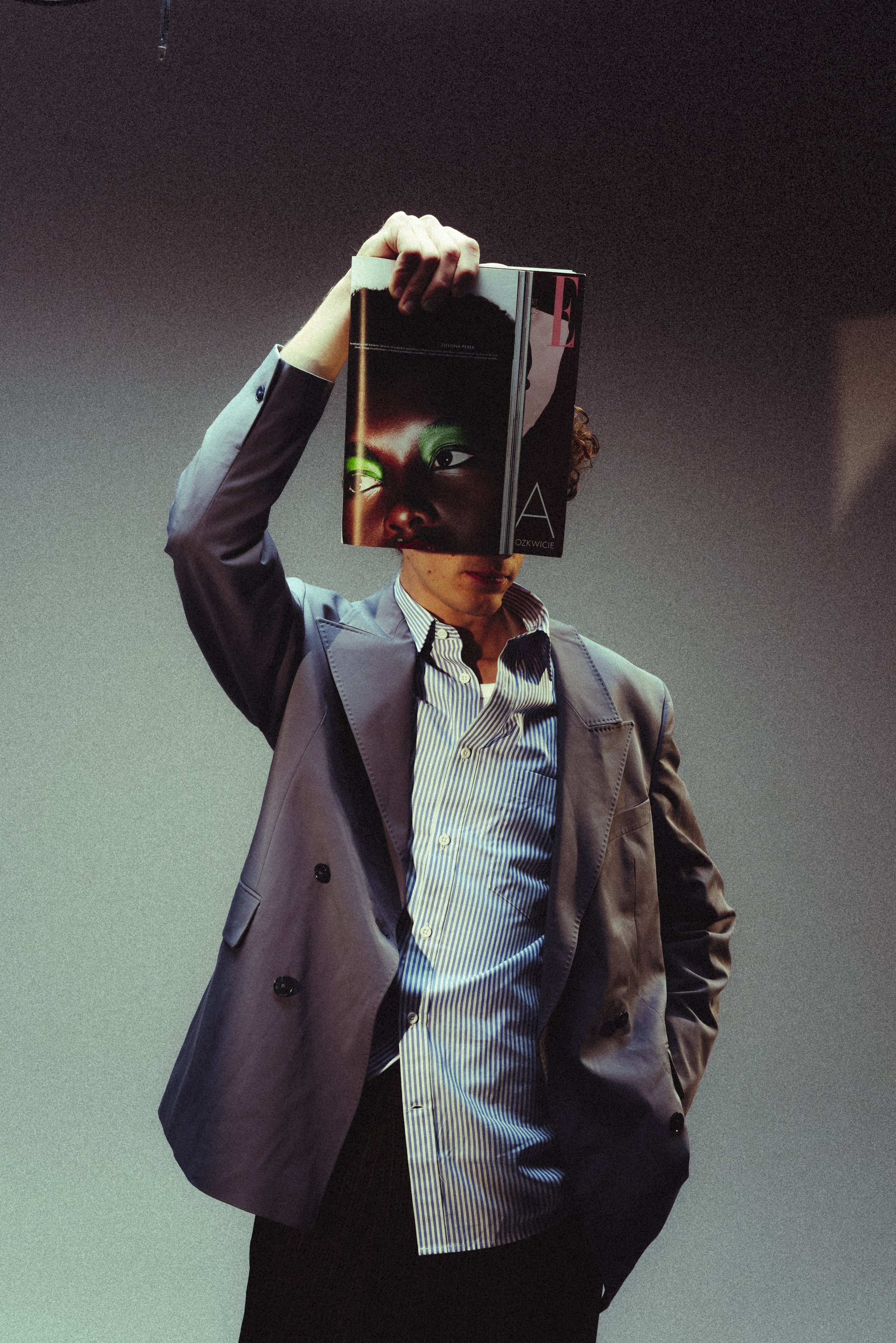 Person standing against a neutral background, wearing a blazer and shirt, holding a magazine with a colorful face on the cover over their face.