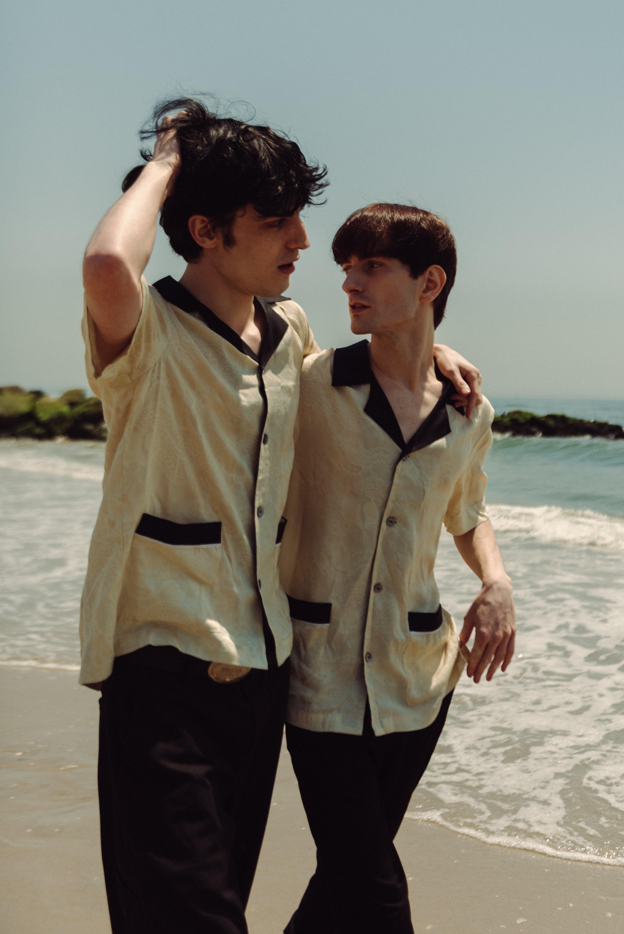 Two young men walking on the beach, wearing matching beige shirts with black trim, with ocean waves and rocks in the background.