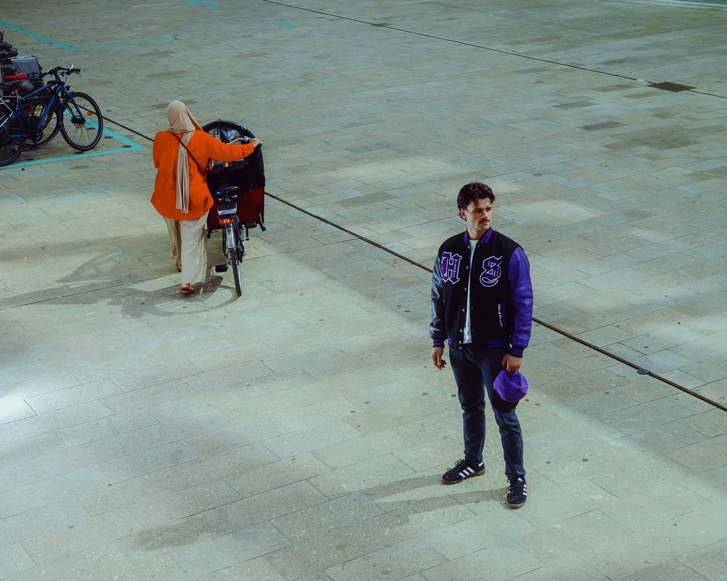 A young man in a black and purple jacket stands on a tiled surface, holding a purple hat, with a woman in an orange jacket and beige pants near a bicycle and a bicycle with a rear cargo rack nearby.