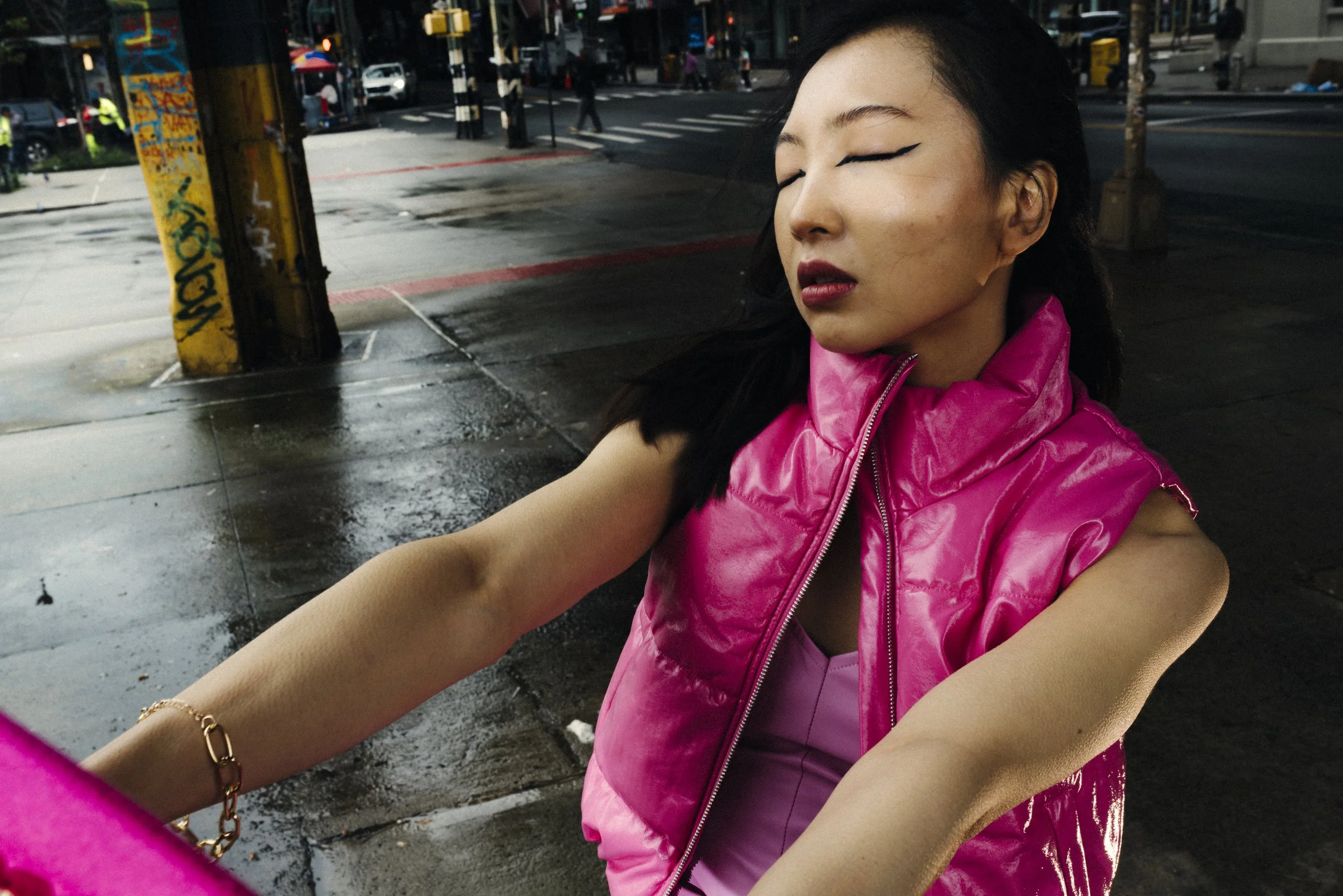 A woman with dark hair and closed eyes wearing a shiny pink vest is taking a selfie on a wet city sidewalk during the daytime, with traffic and pedestrians in the background.