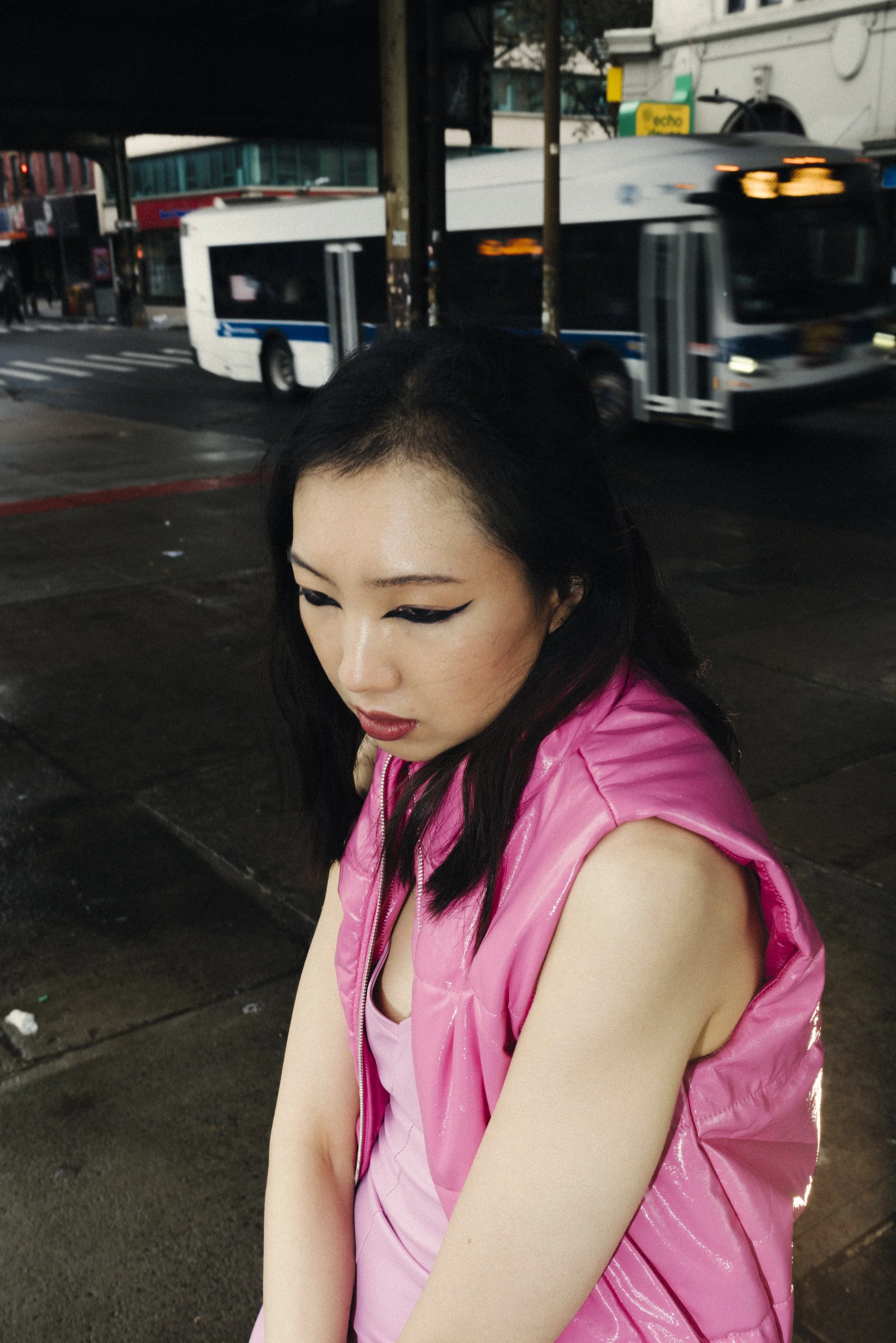 Young woman with black hair and makeup, wearing a pink sleeveless jacket, standing on a city sidewalk with a bus passing by in the background.