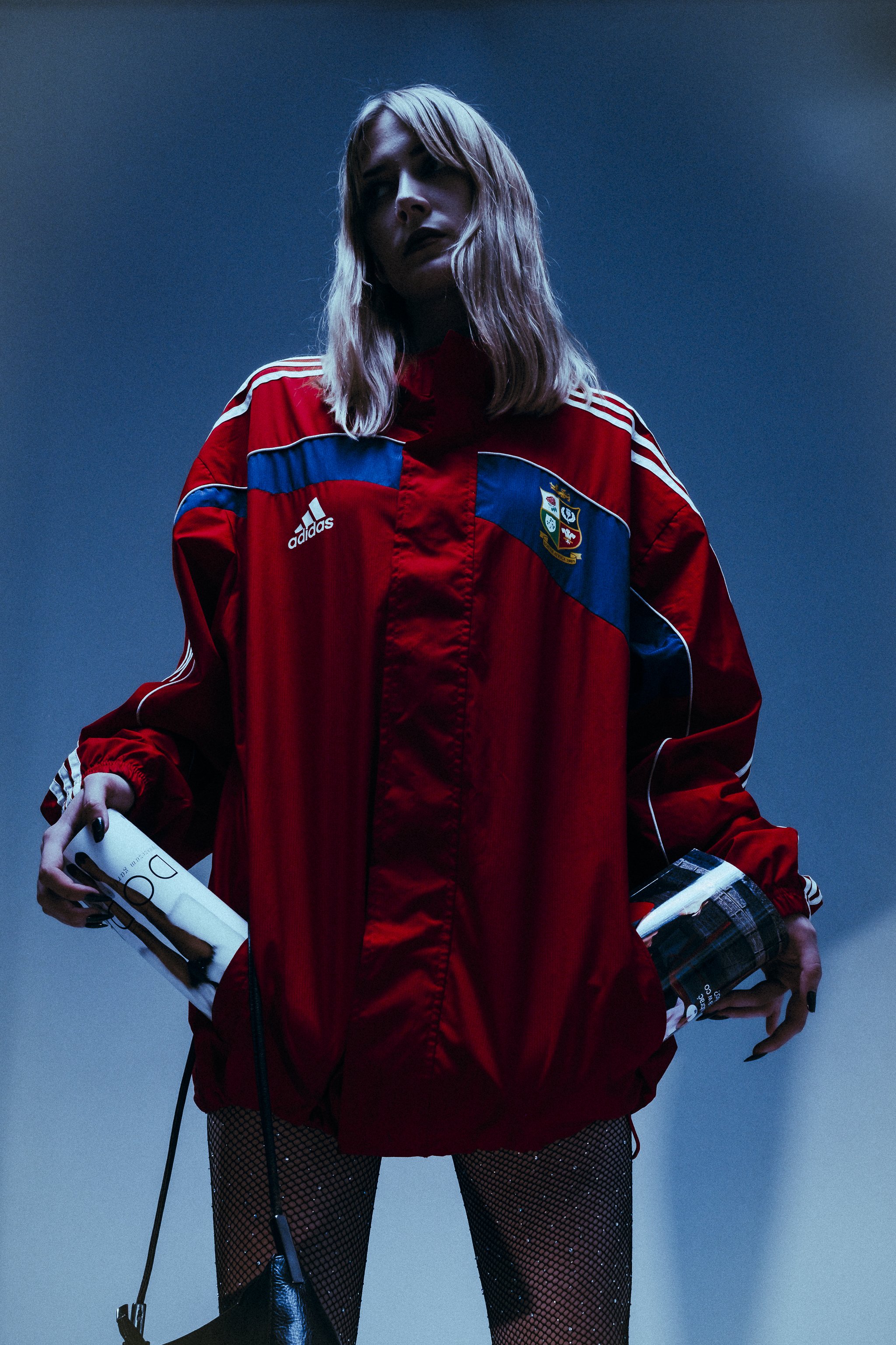 A woman with shoulder-length blonde hair wearing a red Portugal national team Adidas jacket and fishnet stockings, holding a magazine and a small bag, posed against a dark blue background.