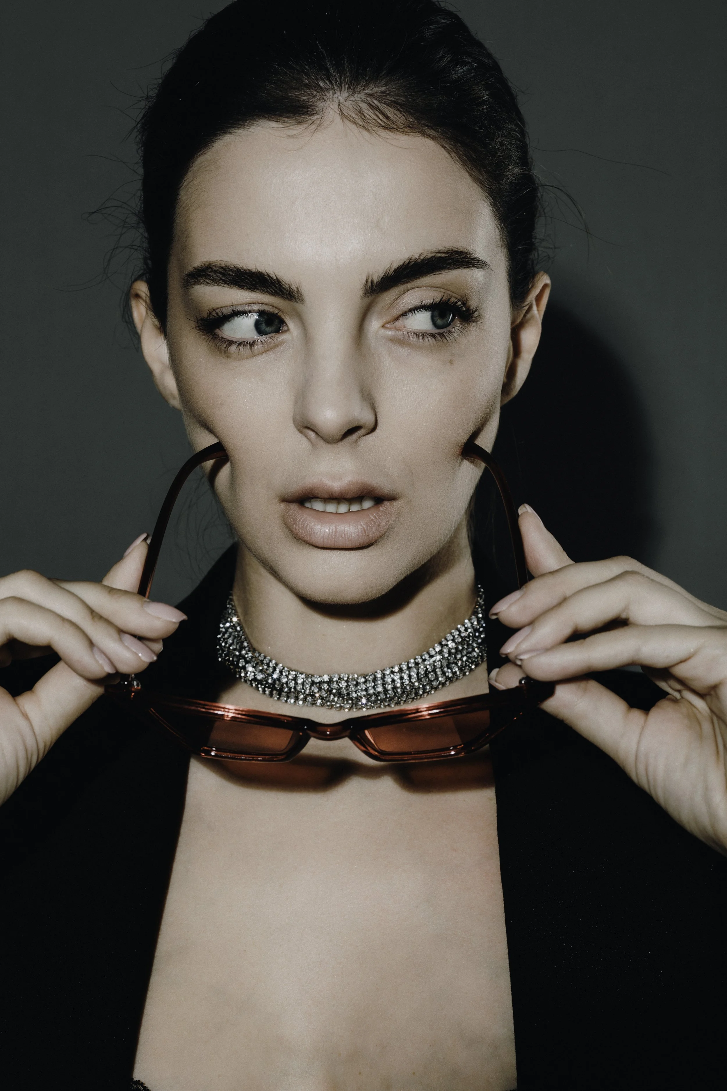 Close-up of a woman with dark hair holding red sunglasses near her face, wearing a sparkling diamond choker necklace, with one eye turned to the side and a neutral background.