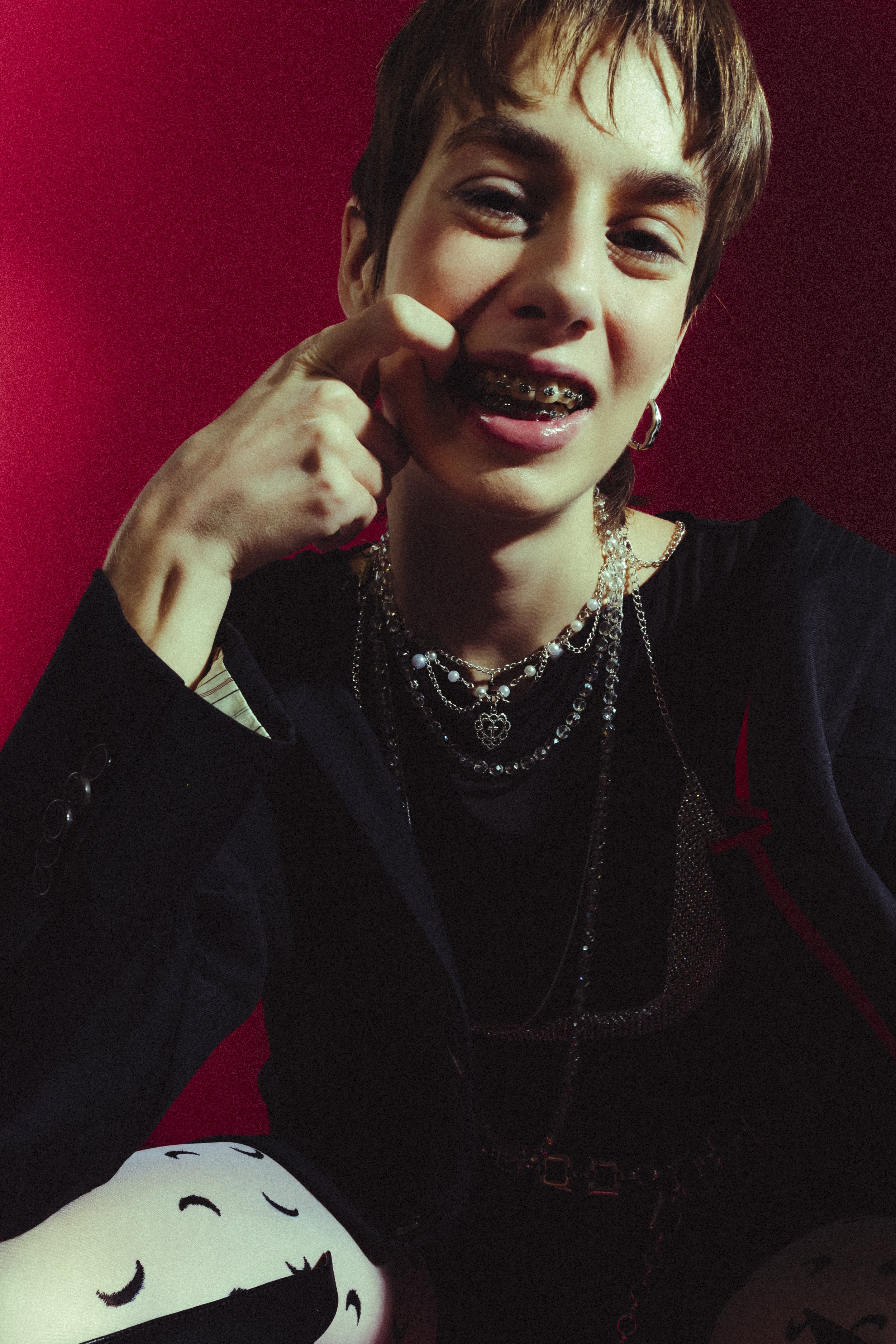 A young person with short hair and multiple layered necklaces, wearing a black jacket, smiling and touching their face, with a red wall background.
