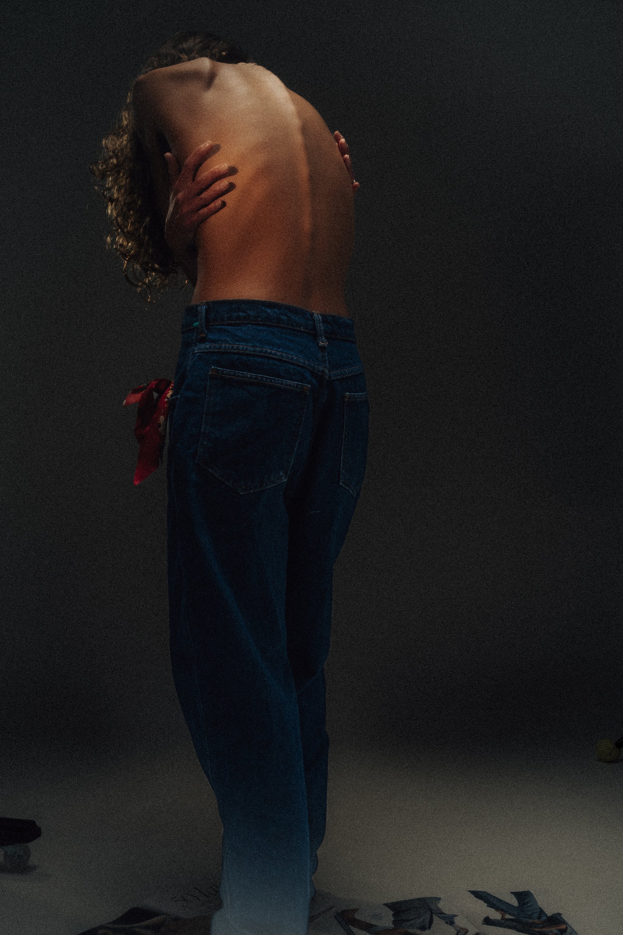 A person with curly hair standing topless with their back to the camera, wearing jeans and a red ribbon tied around the waist, holding their arms crossed over their chest in front of a dark background.
