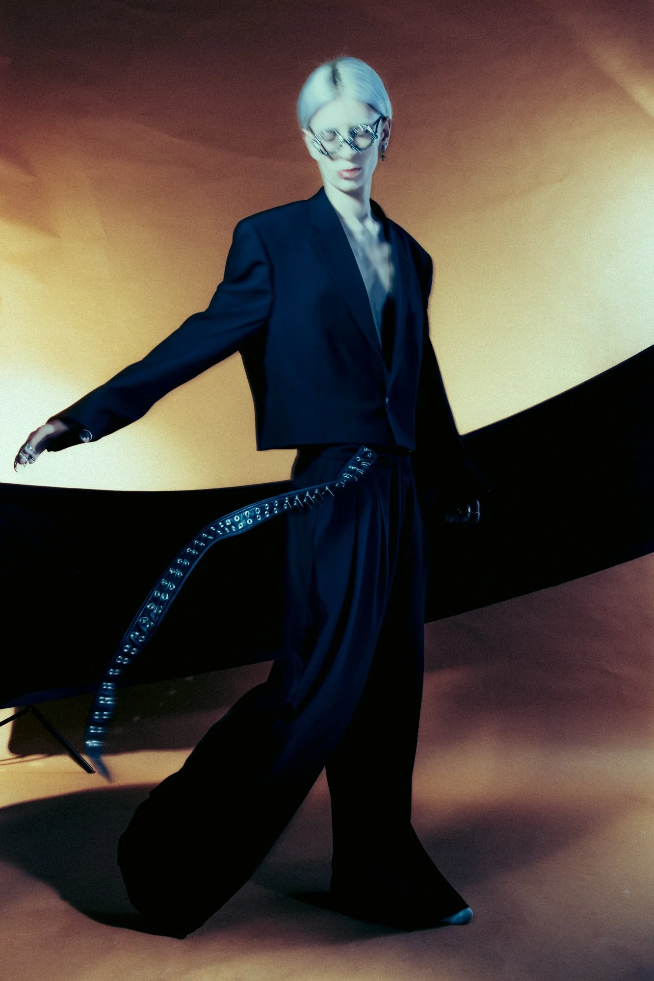 A woman with short blonde hair wearing large sunglasses, a black blazer, and black wide-leg pants. She is standing against an orange background, holding a long black and blue strap or ribbon.