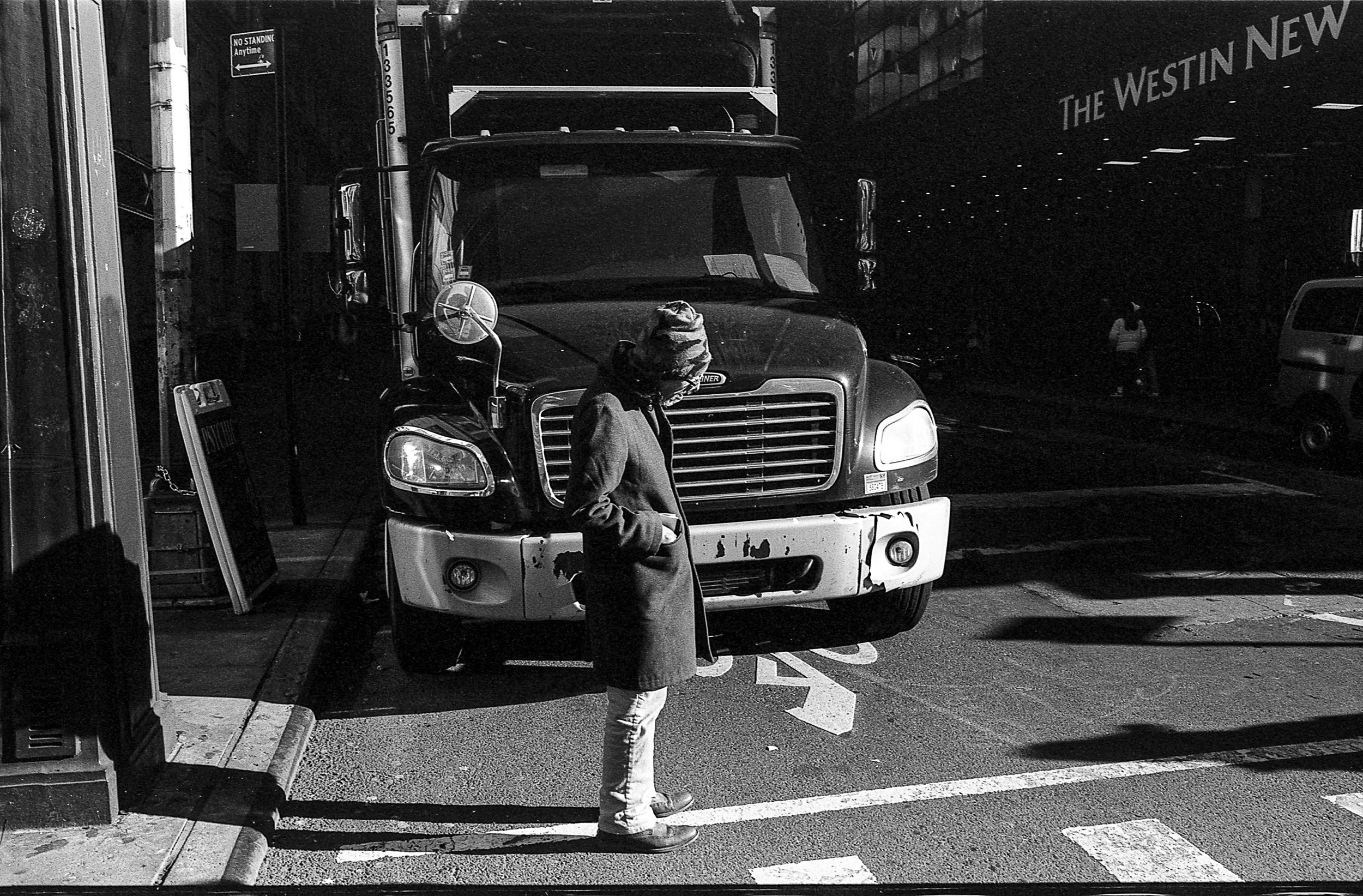 A person wearing a coat and a beanie hat standing on a city street at night, with a large truck parked behind them.