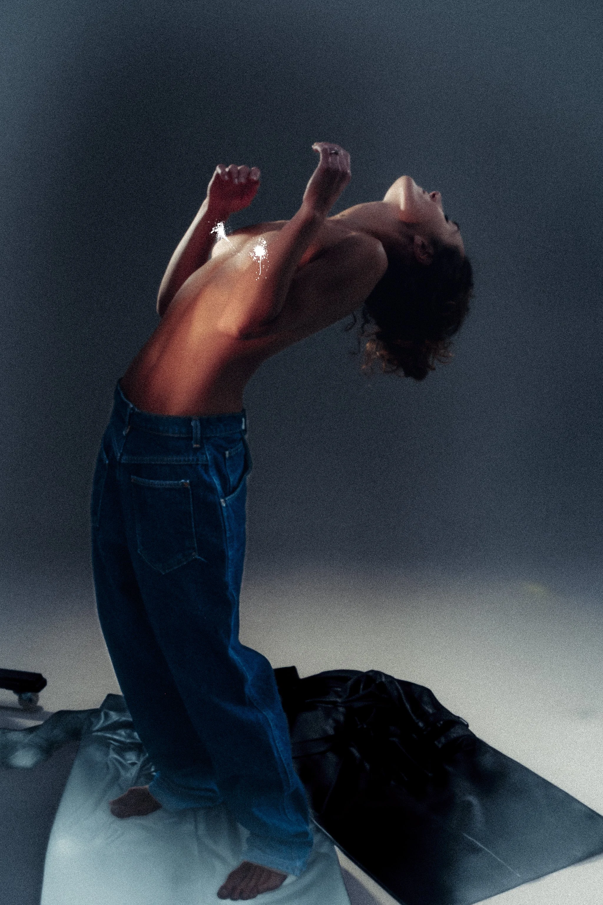 A person standing on an ironing board, leaning backward with their head tilted and mouth open, topless and wearing jeans.