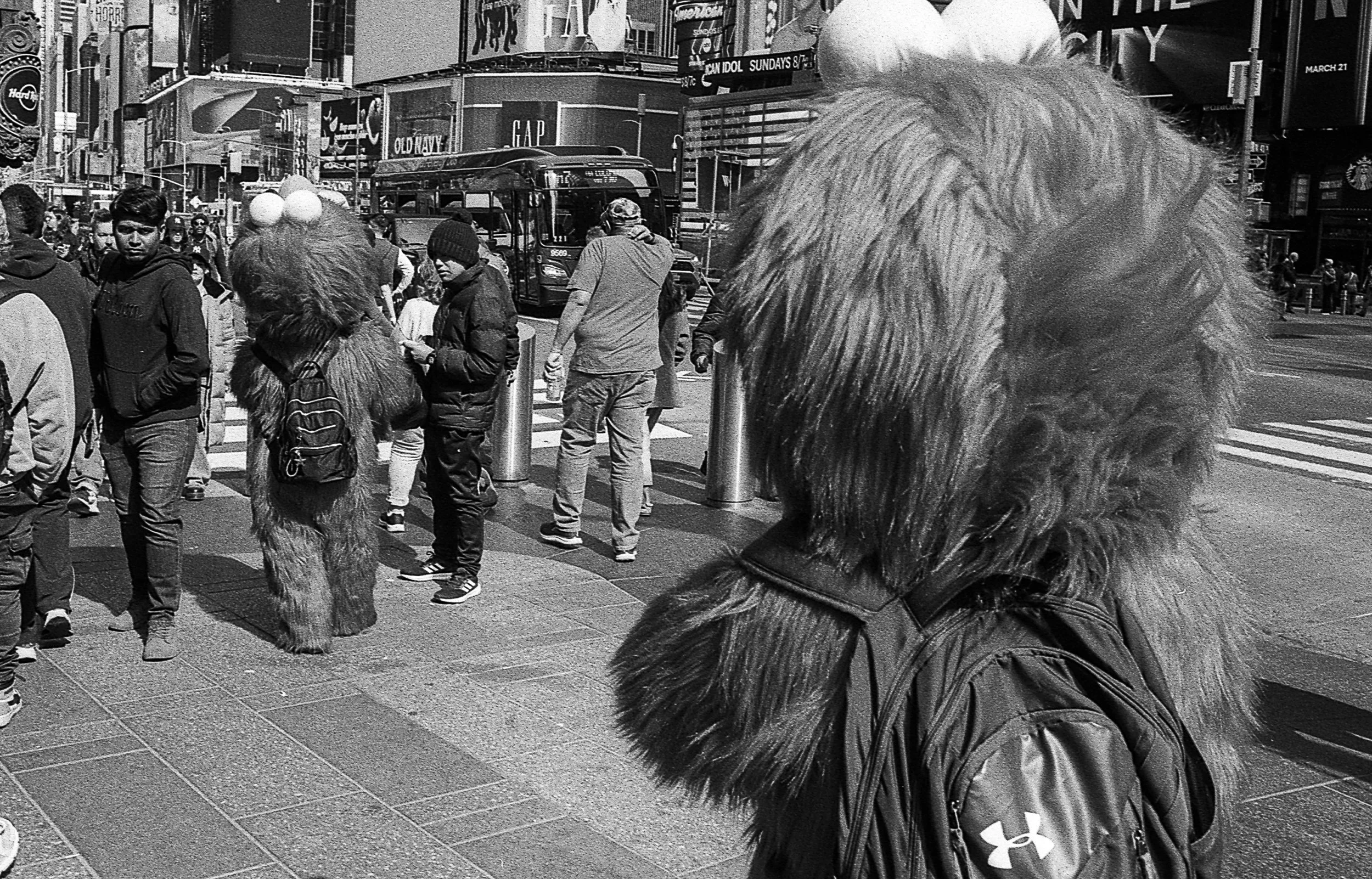 Person in a furry costume with large ears and backpack standing on busy city street, with pedestrians and buildings in the background.