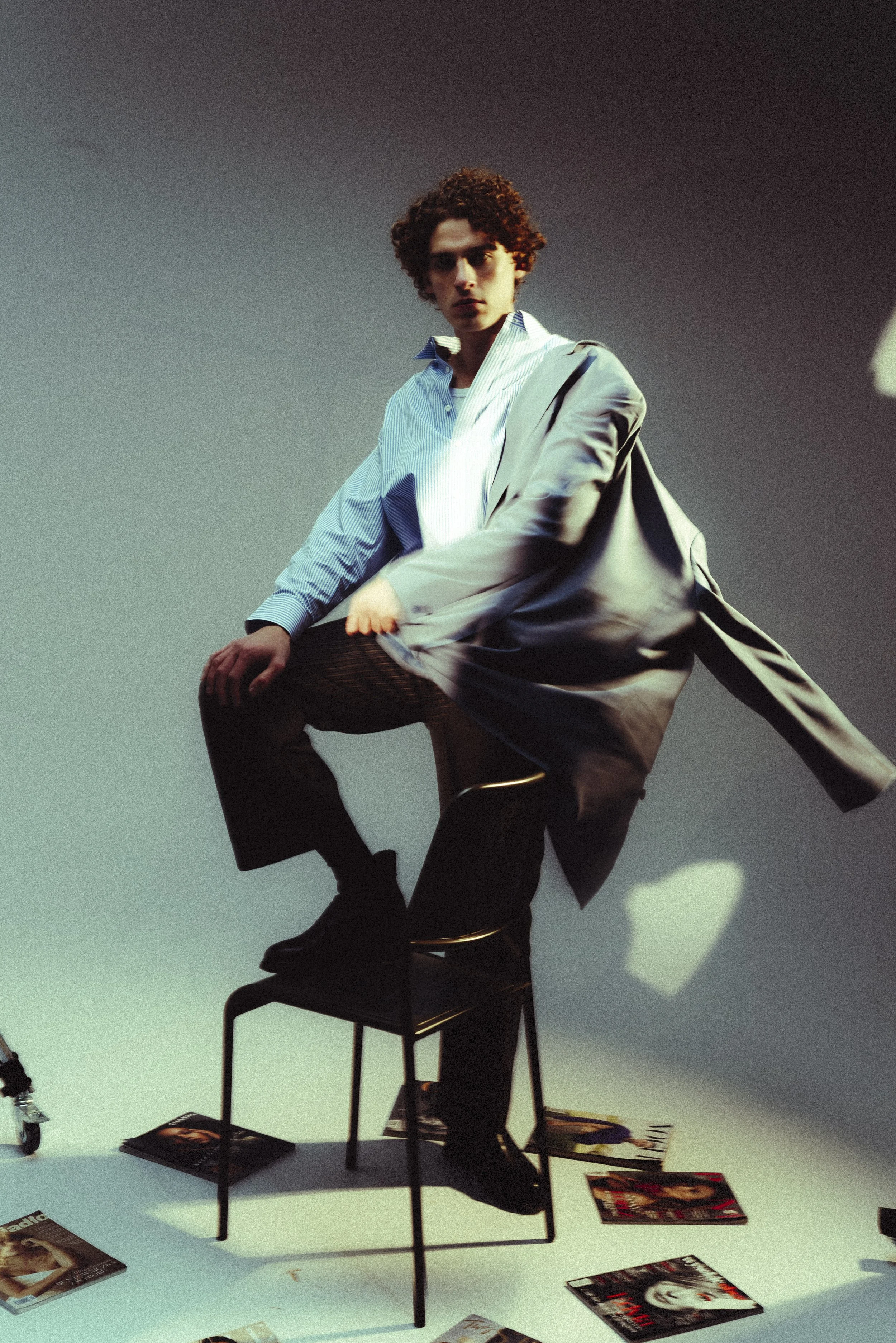 A young man with curly hair sits on a black chair with one leg lifted, wearing a blue striped shirt and a gray coat. He is in a studio with a plain background, surrounded by scattered magazines on the floor.