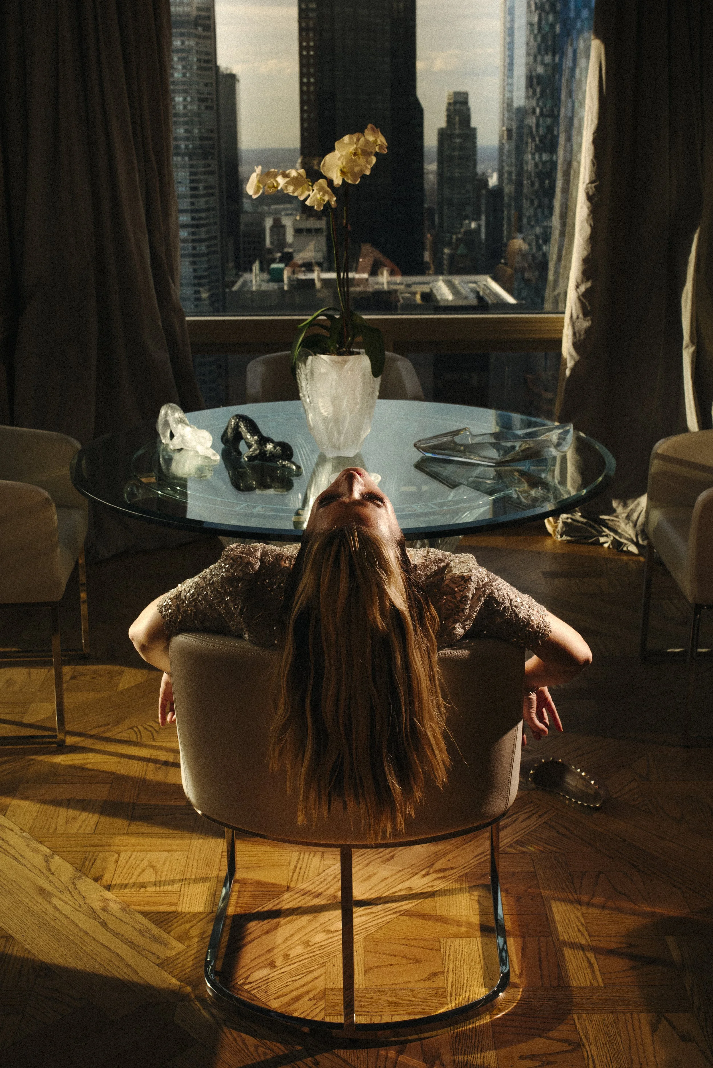 A woman reclining backward in a chair in a modern apartment, with her head and hair hanging over the chair's back, facing a round glass table. The table has a white vase with orchids, some decorative glass sculptures, and a pair of glasses. Large win