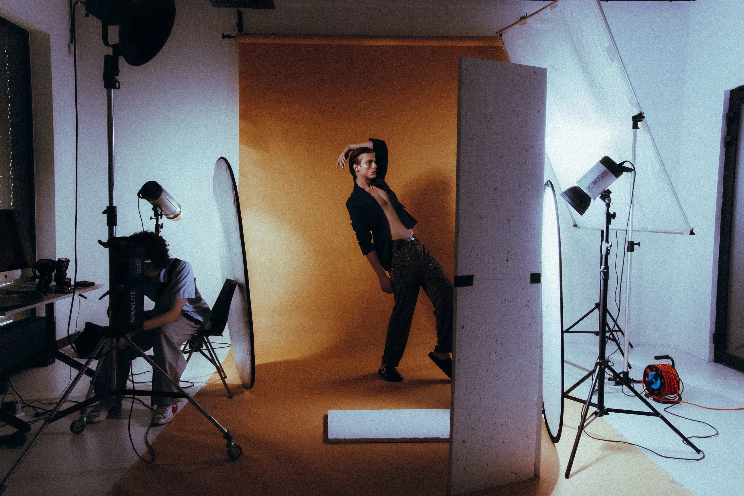 A male model posing in a photography studio with a yellow backdrop, surrounded by lighting and photo equipment, while a photographer adjusts a camera.