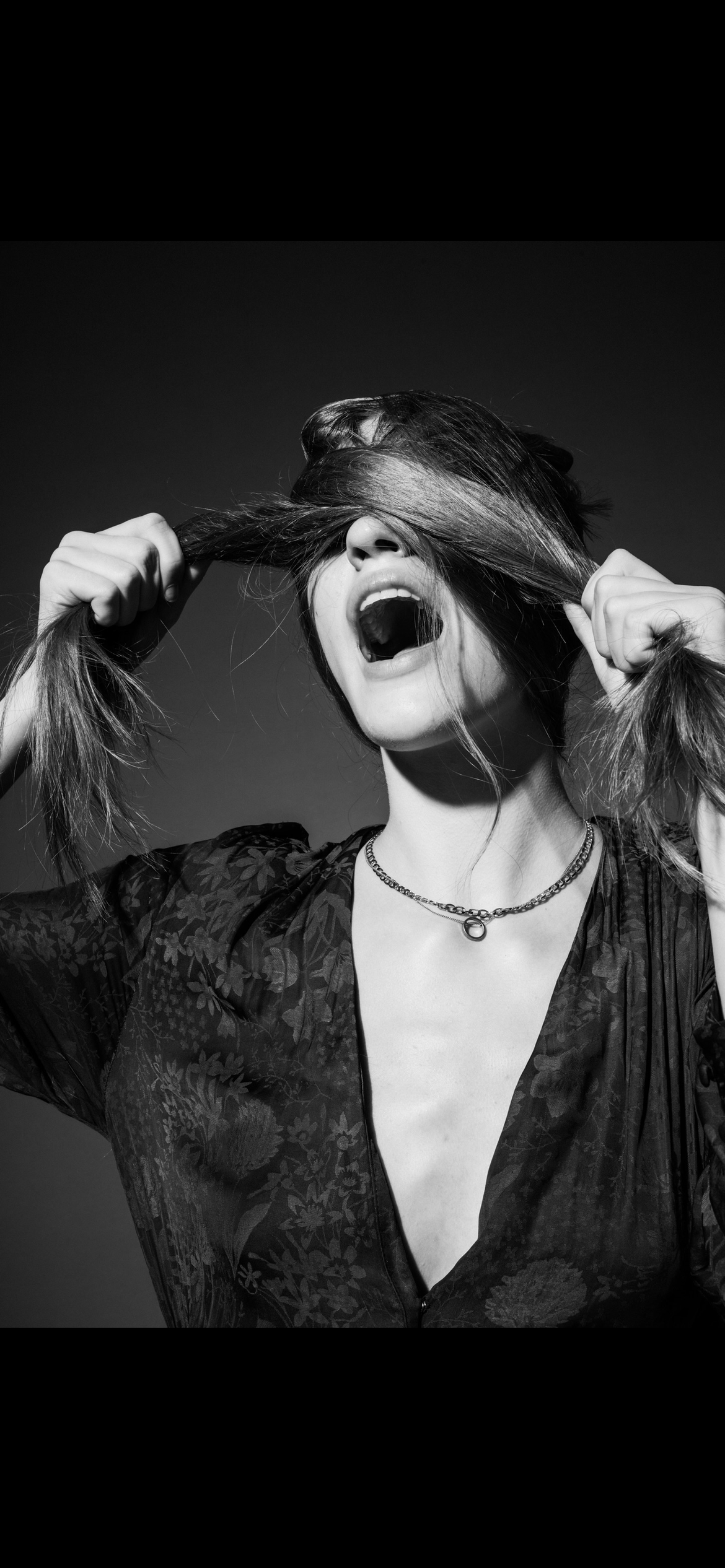 Black and white photo of a woman with long hair, holding her hair over her eyes, with her mouth open as if shouting.