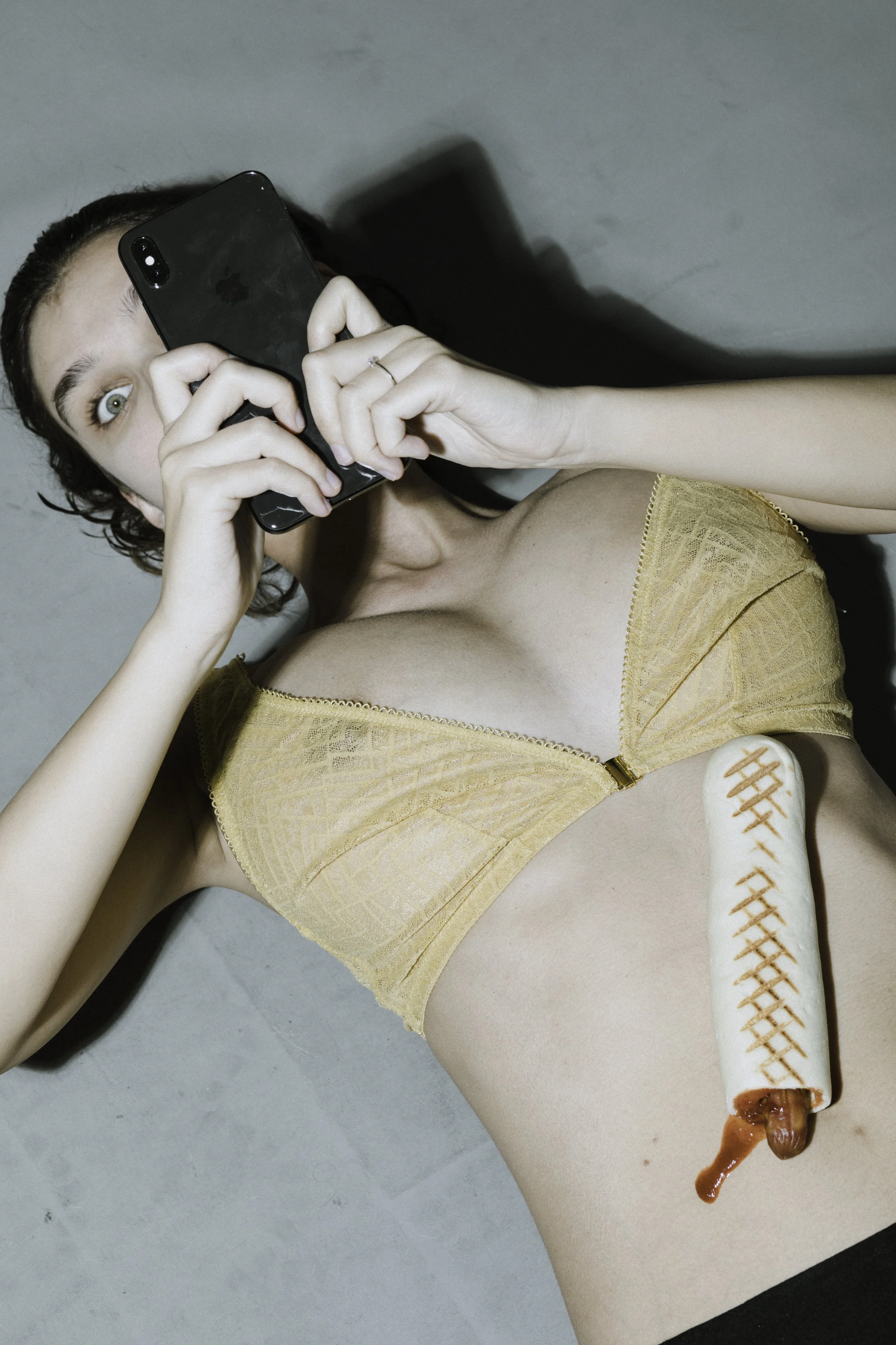 Young woman lying on her back taking a selfie with a smartphone, wearing a yellow bra, with a large novelty hot dog prop placed on her torso.