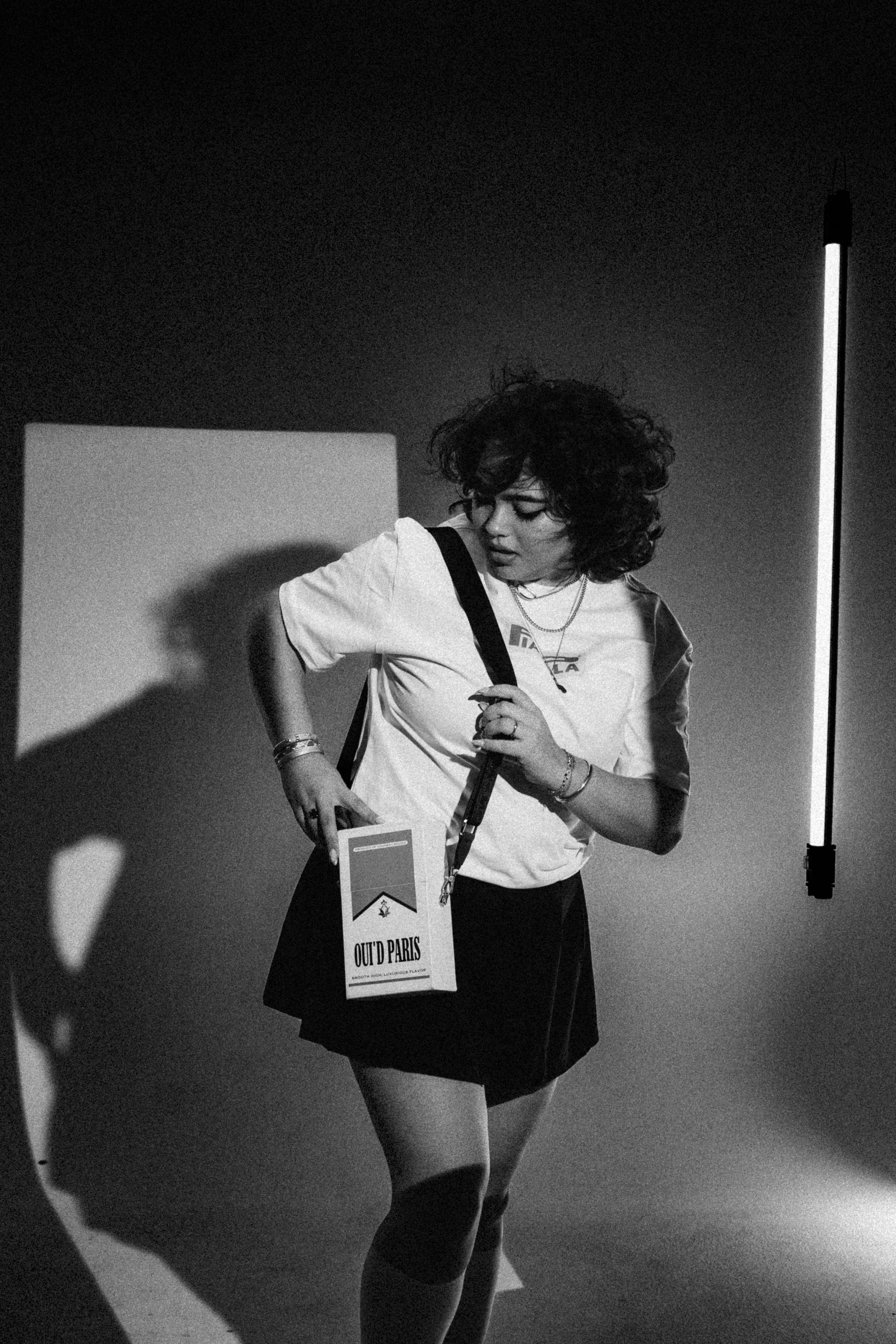 A woman with curly hair in a black and white photo, wearing a white T-shirt, black skirt, and knee-high socks, standing against a plain background with a vertical light source. She has several accessories, including rings, bracelets, and necklaces, a