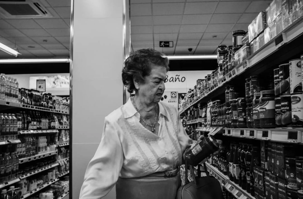 An elderly woman shopping in a supermarket aisle, examining a bottle of product on the shelf.