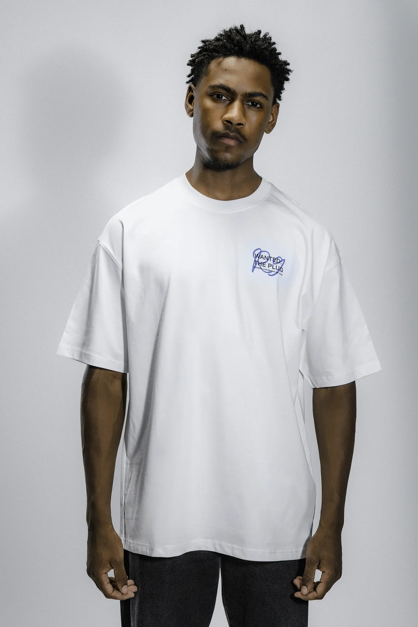A young man with short, curly black hair and a serious expression, wearing a plain white T-shirt with a small blue graphic on the chest that reads "Wanted The Plug" and dark pants, standing against a plain light grey background.