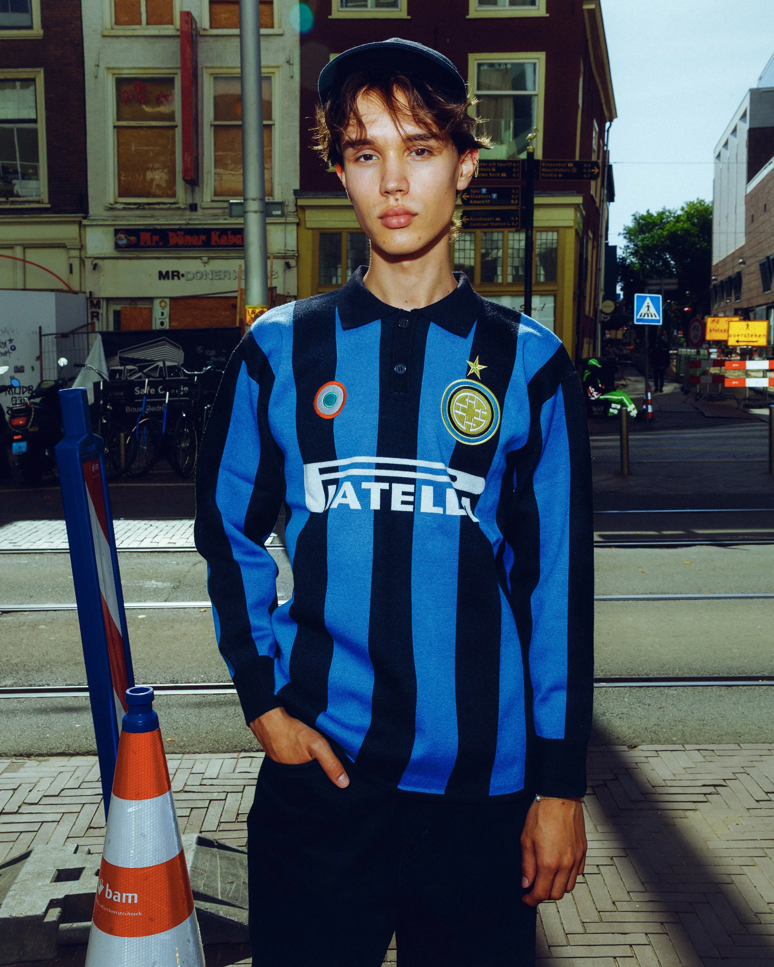A young man in an Inter Milan football jersey standing outdoors on a city street, with buildings, street signs, and tram tracks visible in the background.