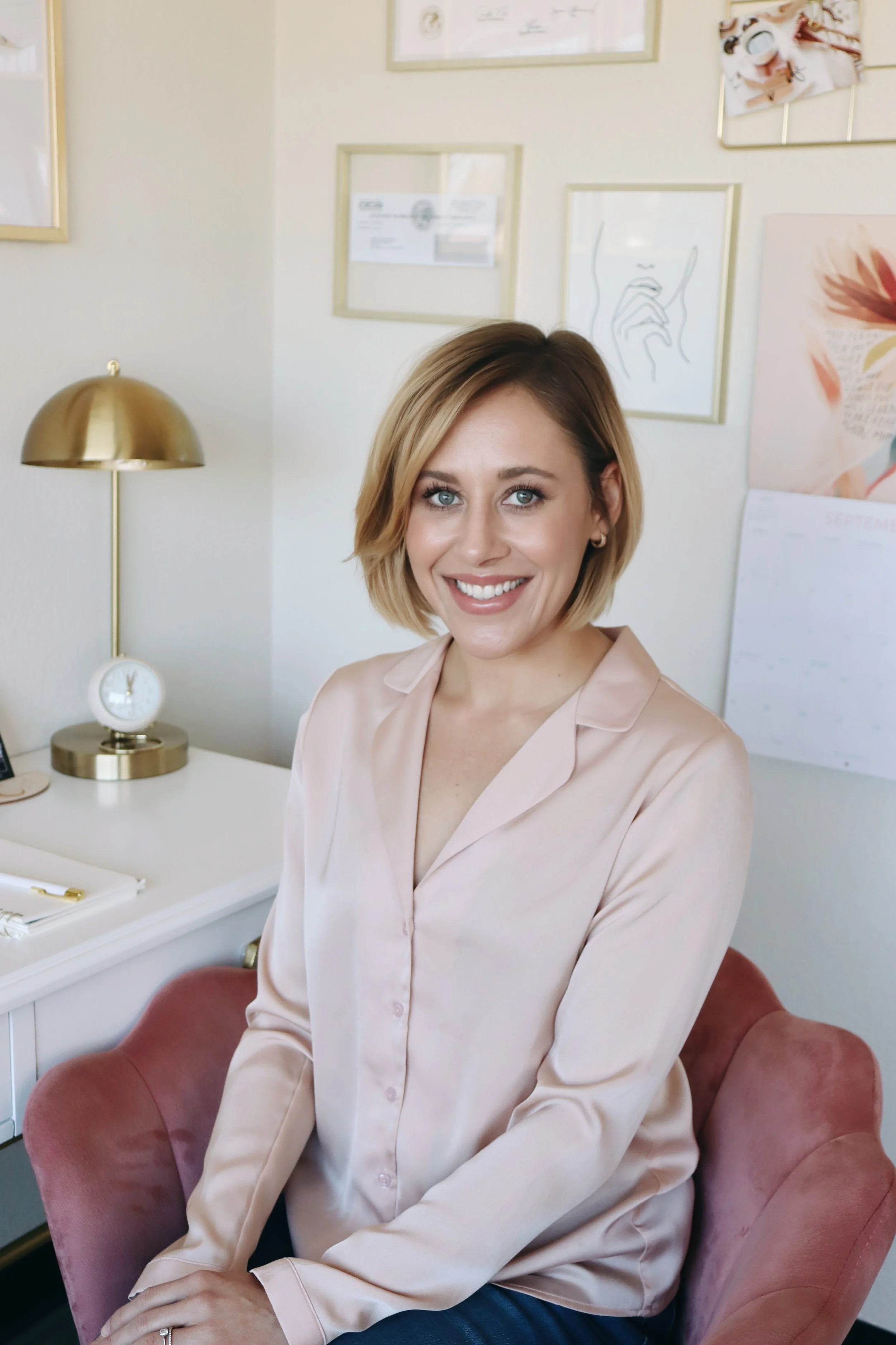 A woman with short blonde hair and blue eyes smiling while sitting on a pink chair in a cozy, well-decorated room.