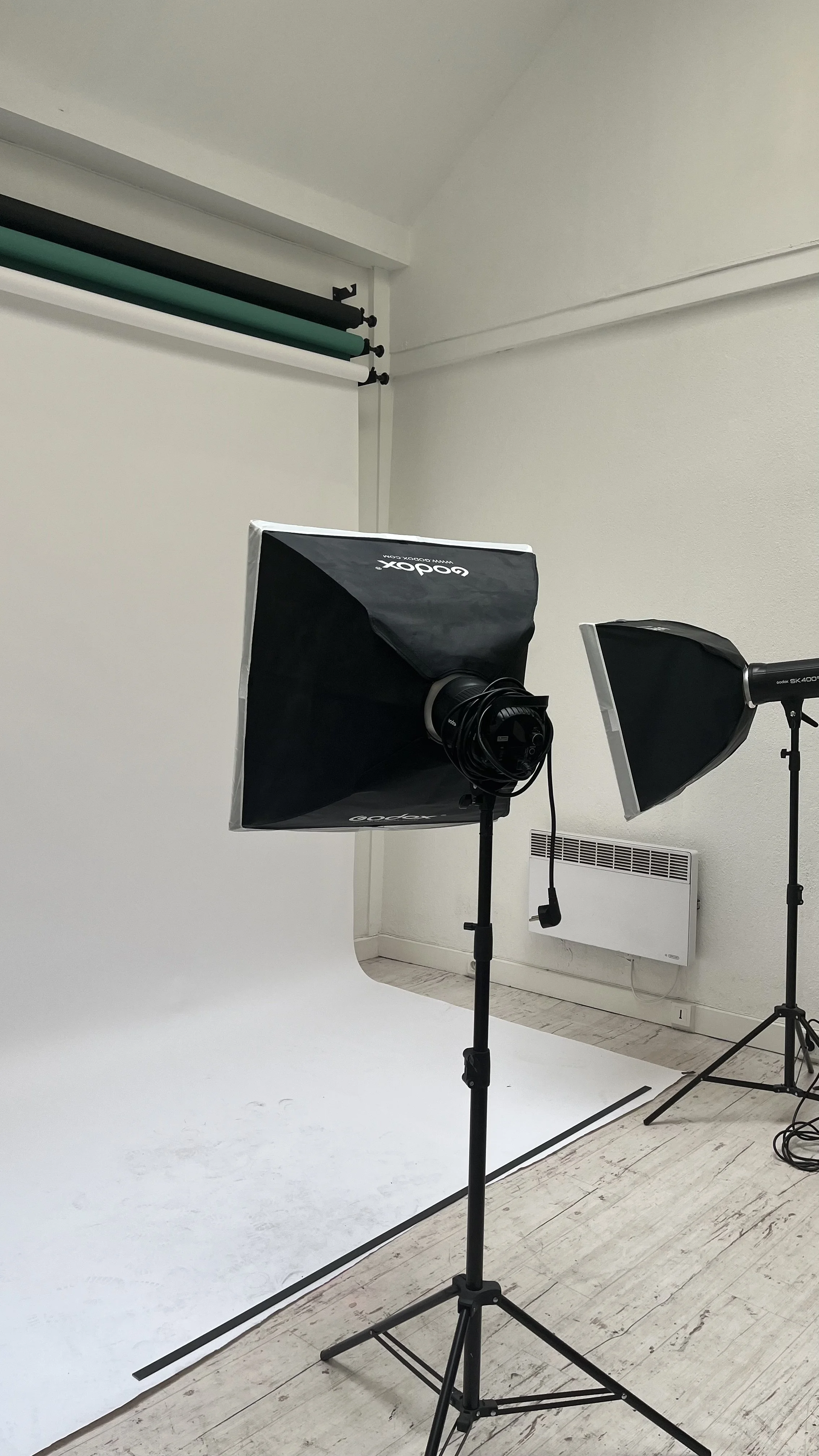Photo of a photography studio with two studio lights on stands, a white backdrop, and a white wall with a heater.