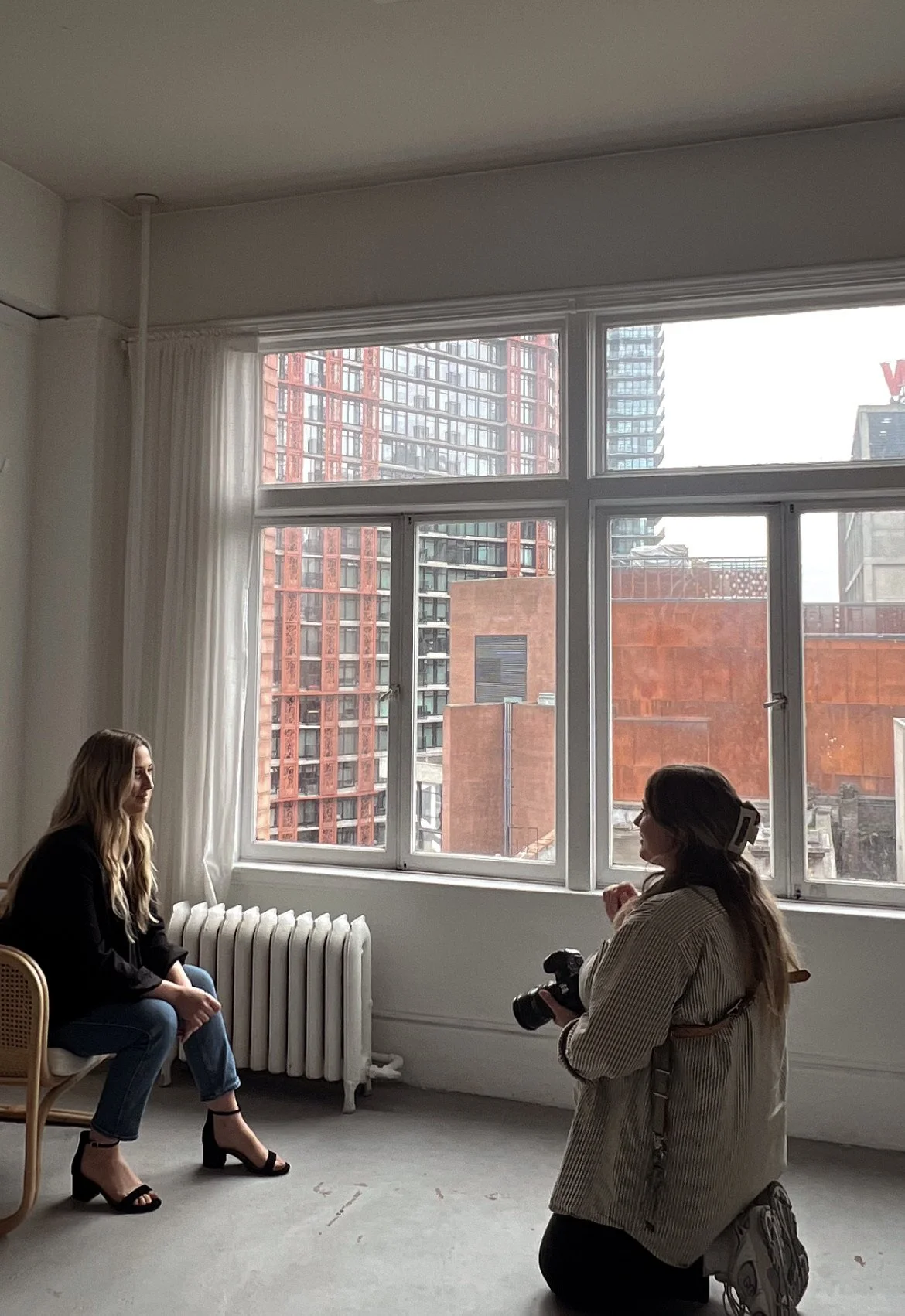 A woman sitting on a chair being photographed by a photographer holding a camera inside a bright room with large windows showing an urban cityscape.