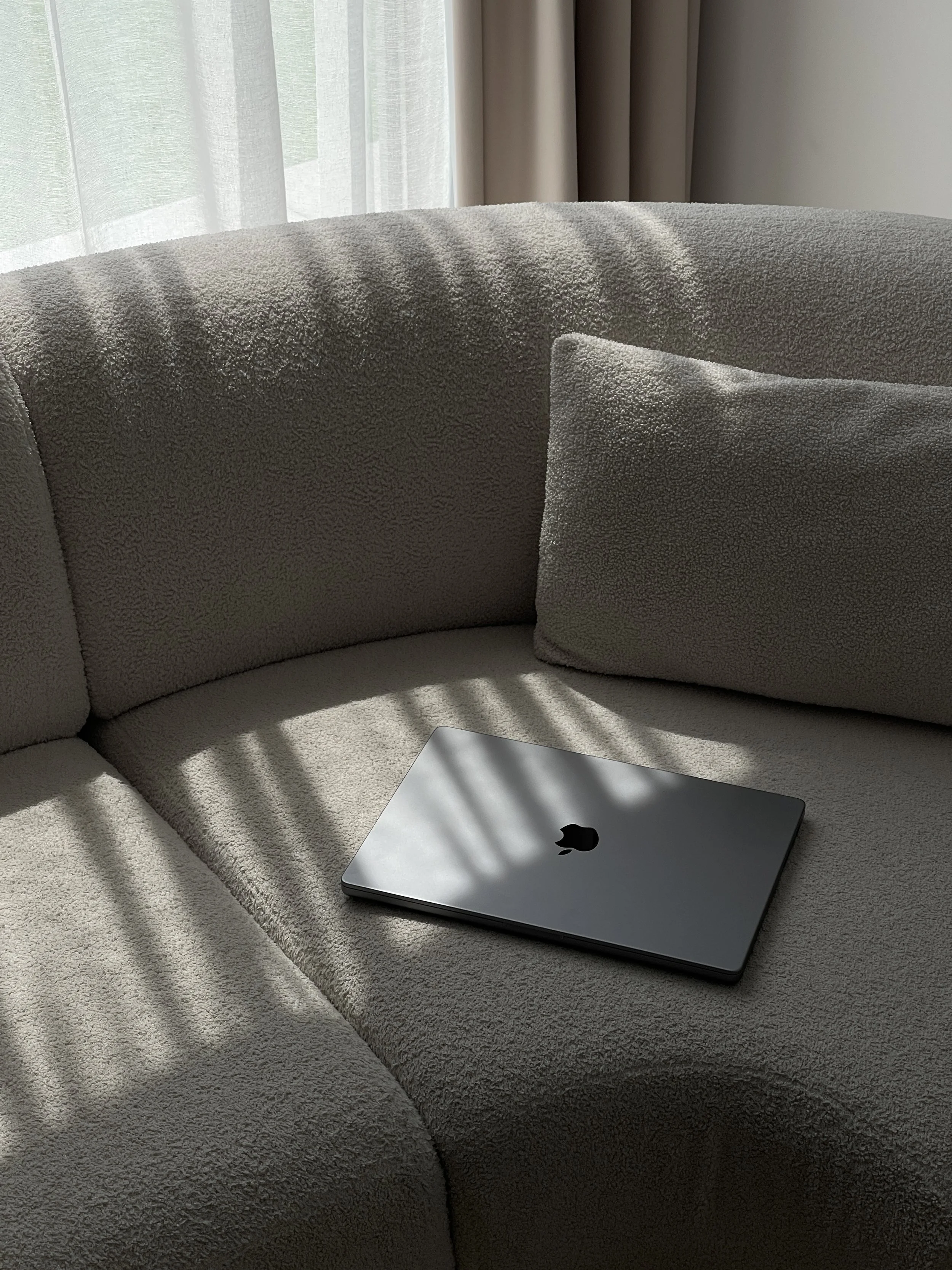 A closed silver MacBook laptop on a beige upholstered couch with pillow, sunlight coming through sheer curtains and casting shadows.