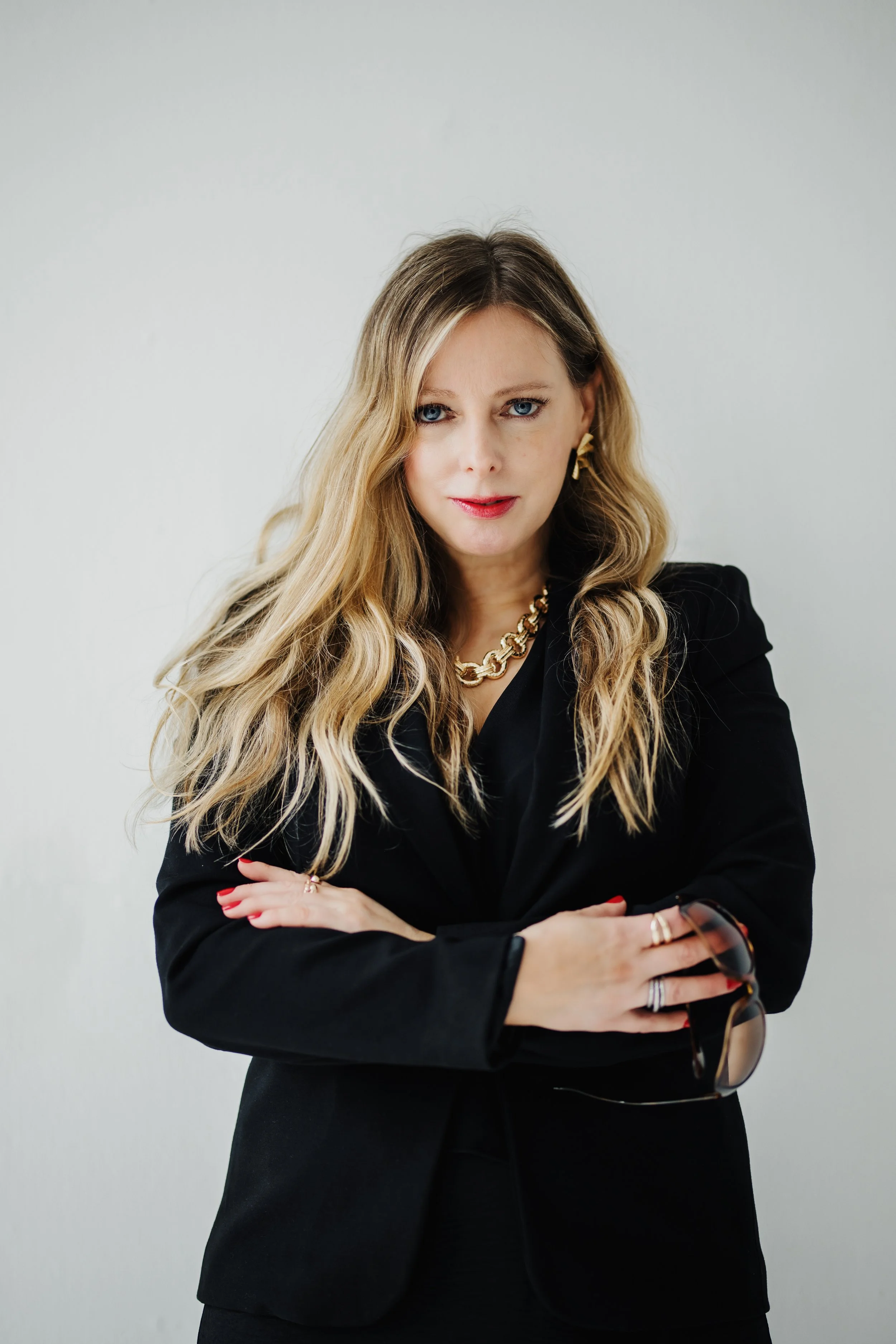 A woman with blonde wavy hair wearing a black blazer and gold jewelry, holding sunglasses, standing against a plain light background.