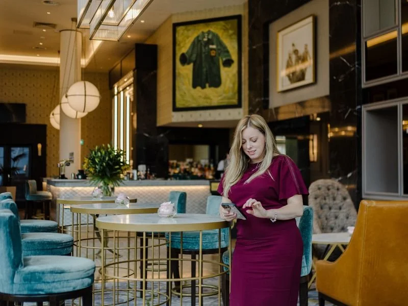 A woman in a maroon dress standing in a stylish hotel lobby, looking at her phone, surrounded by modern furniture and artwork.