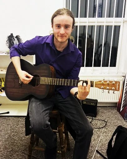 A young man with long hair, wearing a purple shirt, sitting on a wooden stool, holding an acoustic guitar in a room with window bars, a small amplifier, and a music stand.