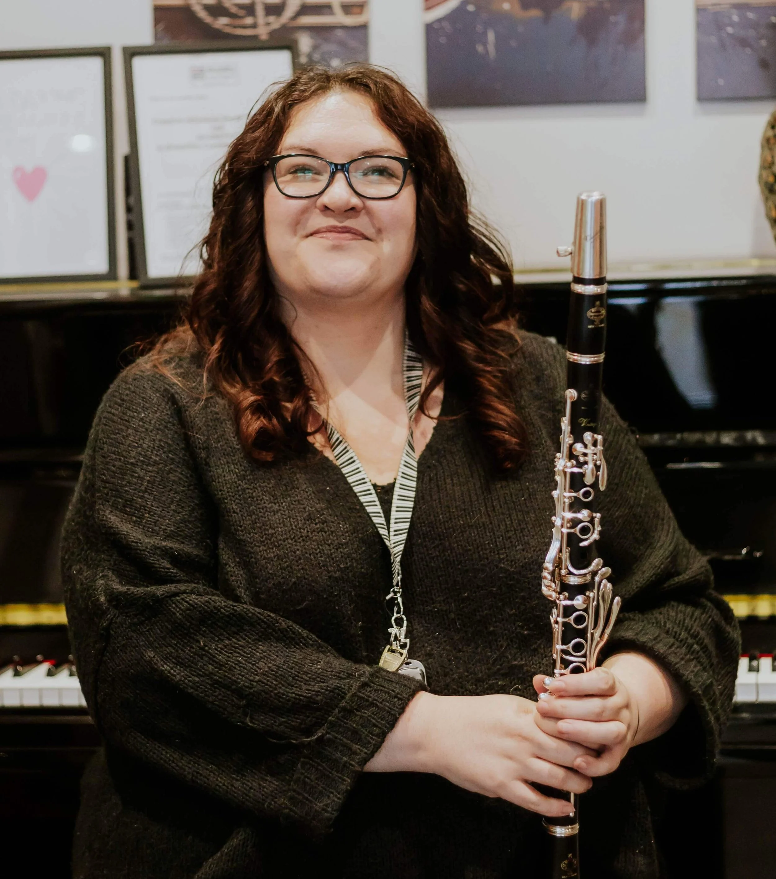 Woman with glasses holding a clarinet in front of a black piano and framed pictures on the wall.