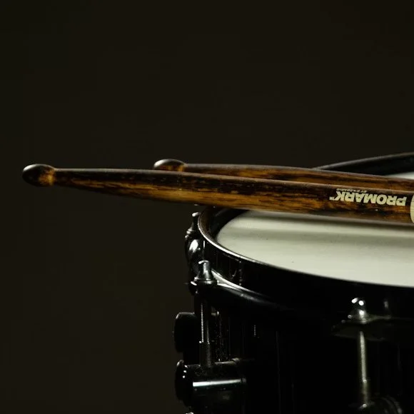 Close-up of a snare drum with a pair of wooden drumsticks resting on it, against a dark background.
