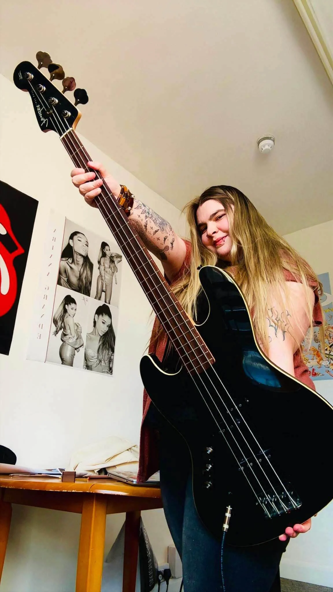 A young woman with long blonde hair holding a black acoustic guitar in a room decorated with black and white posters of women on the wall.