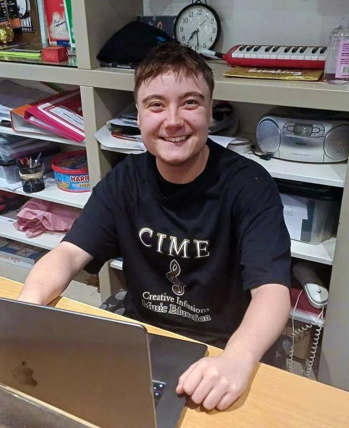 Young man sitting at a desk with a laptop, smiling at the camera, with shelves in the background containing books, files, and a clock.