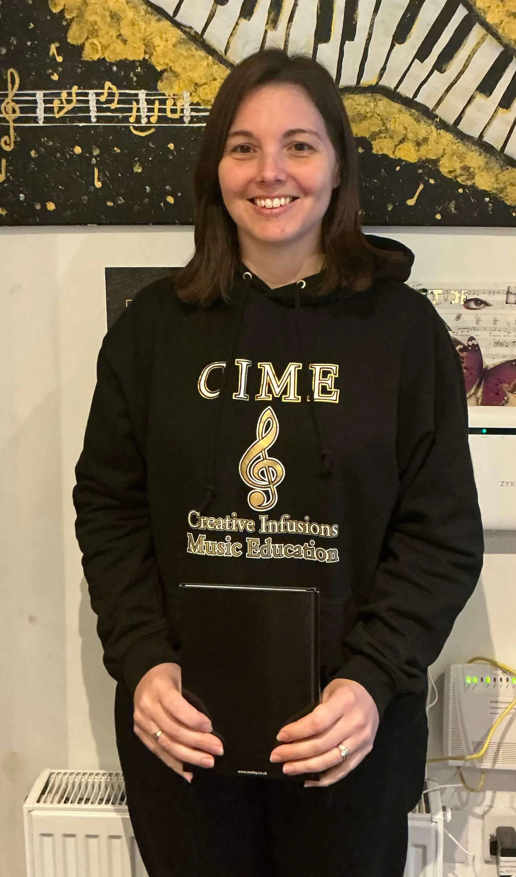 A woman standing indoors, wearing a black hoodie with the words 'CIME' and 'Creative Infusions Music Education' printed on it, smiling, holding a black book, with a musical-themed painting in the background.