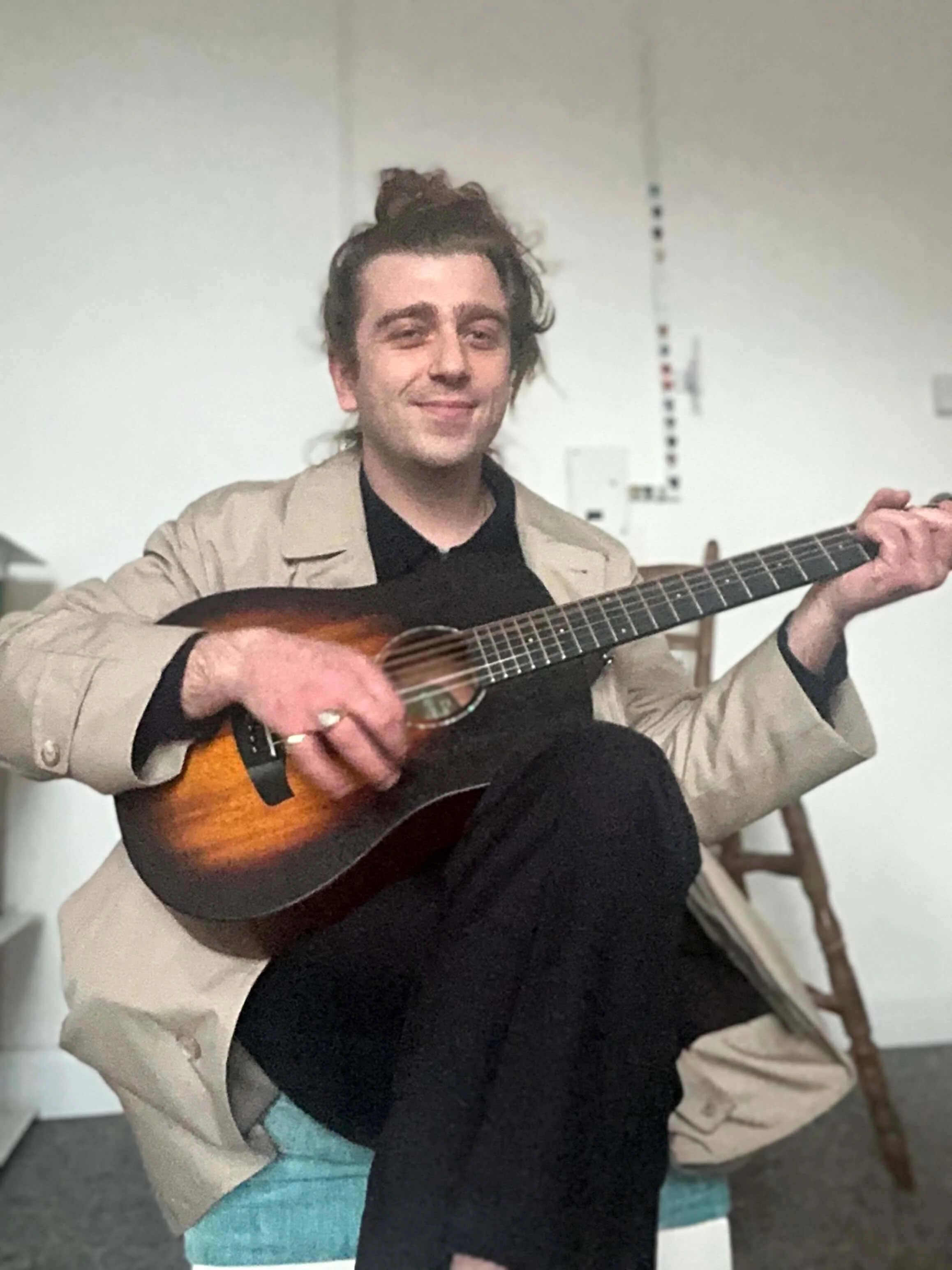 A person with brown hair tied in a bun, playing an acoustic guitar, wearing a beige jacket over a black shirt, sitting on a blue cushion, with a beige blanket draped over their lap, in a room with a white wall and a wooden chair in the background.