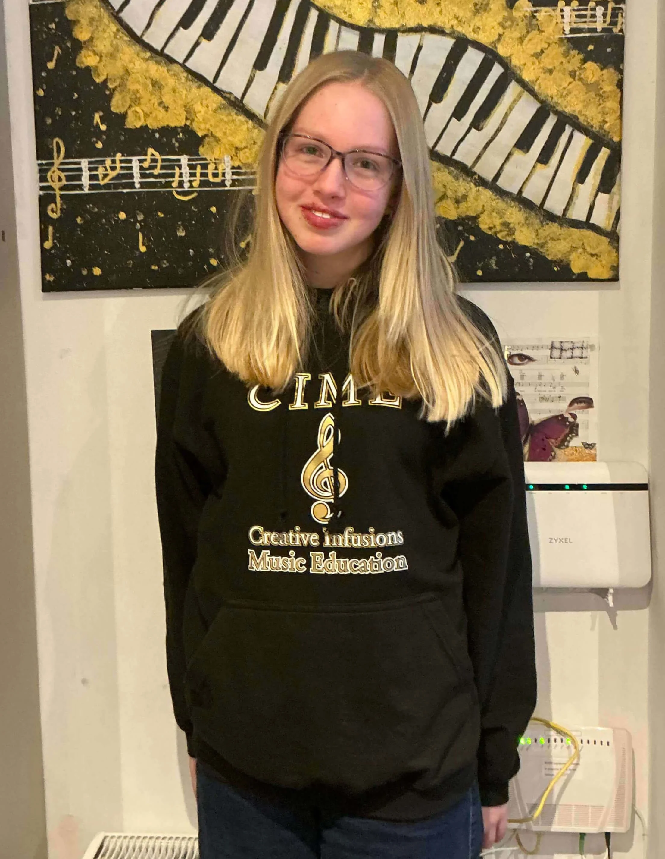 A woman with glasses and long blonde hair standing in front of a wall with art and musical symbols. She is wearing a black sweatshirt with gold lettering and a treble clef symbol. The background includes a painting of a piano keyboard with yellow accents and sheet music.