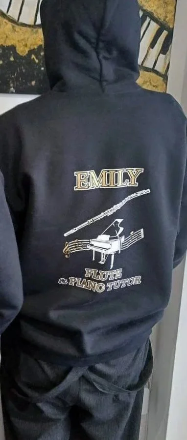 Back of a person wearing a black hoodie with a design that includes a flute and a grand piano, with the words "Emily" and "Flute & Piano Tutor."