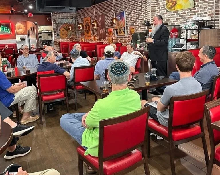 Group of people seated around tables in a restaurant listening to a man standing and speaking. The restaurant has red chairs, brick walls, wall art, and a ceiling fan.