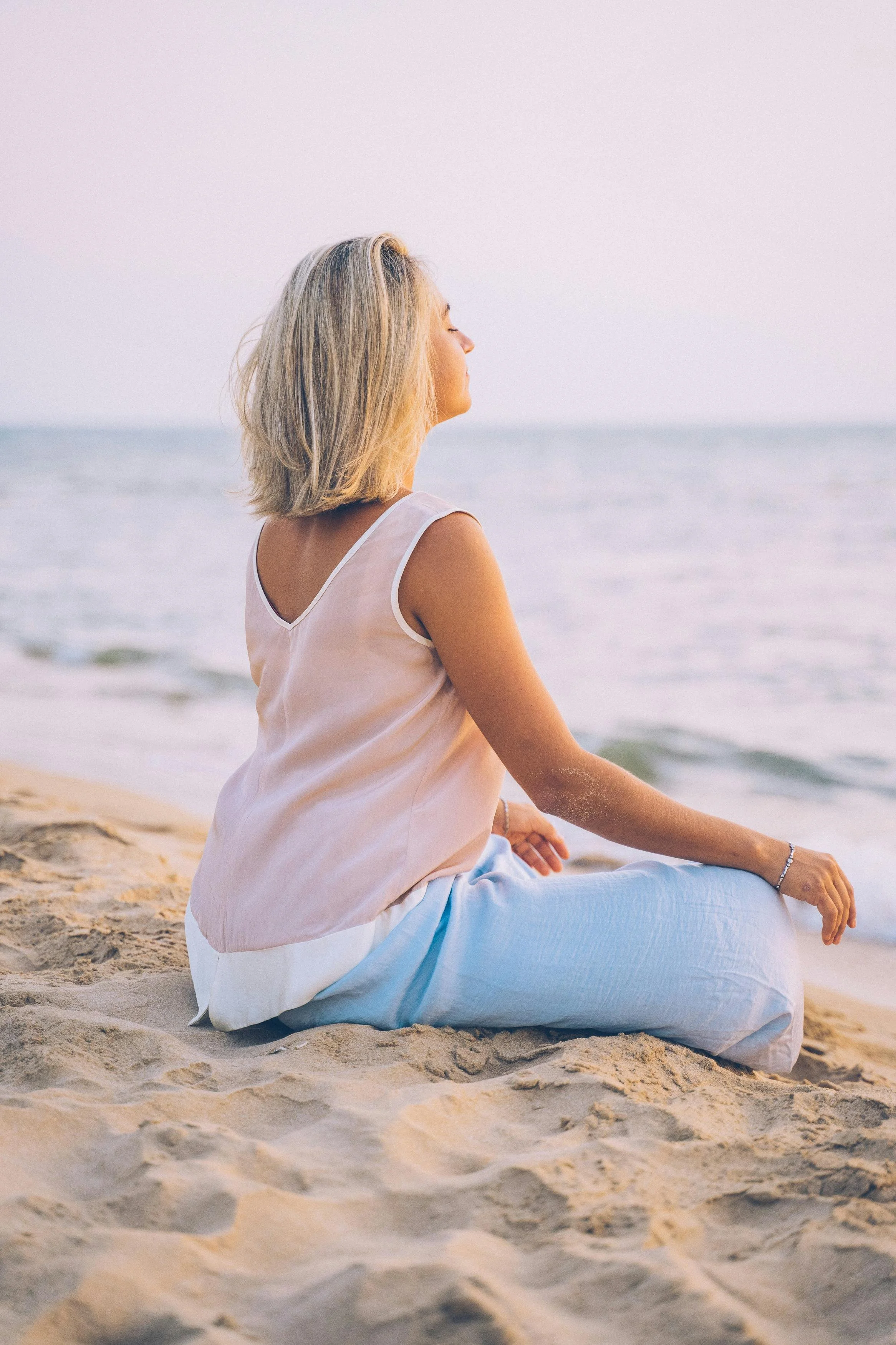 Woman sitting on the sand at the beach, facing the ocean with her eyes closed, possibly meditating to reduce anxiety