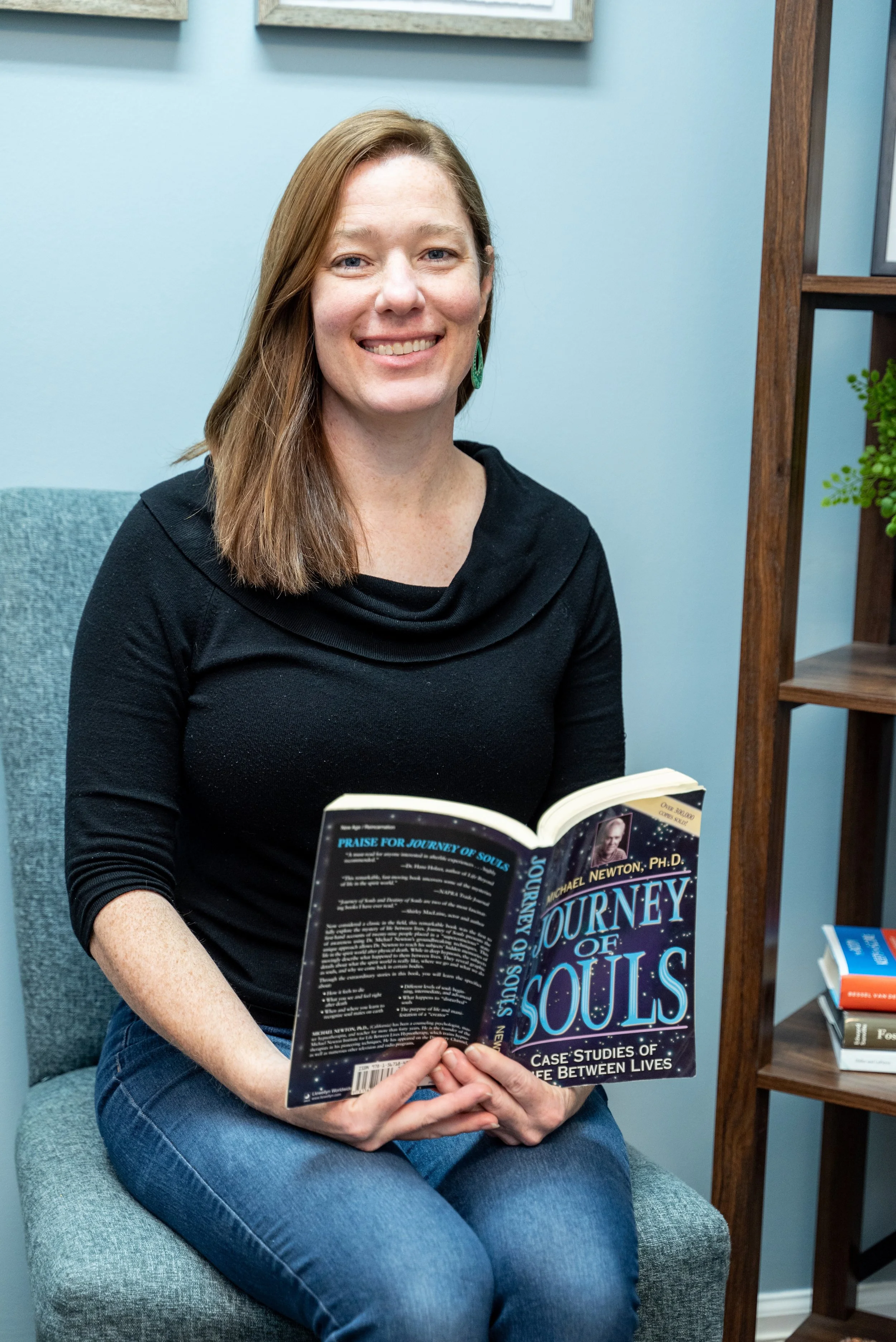 Certified Life Between Lives hypnotherapist and hypnotherapy certification instructor Jennifer Catlin is sitting in a chair holding the book Journey of Souls by Dr. Michael Newton which is a book about Life Between Lives Hypnotherapy