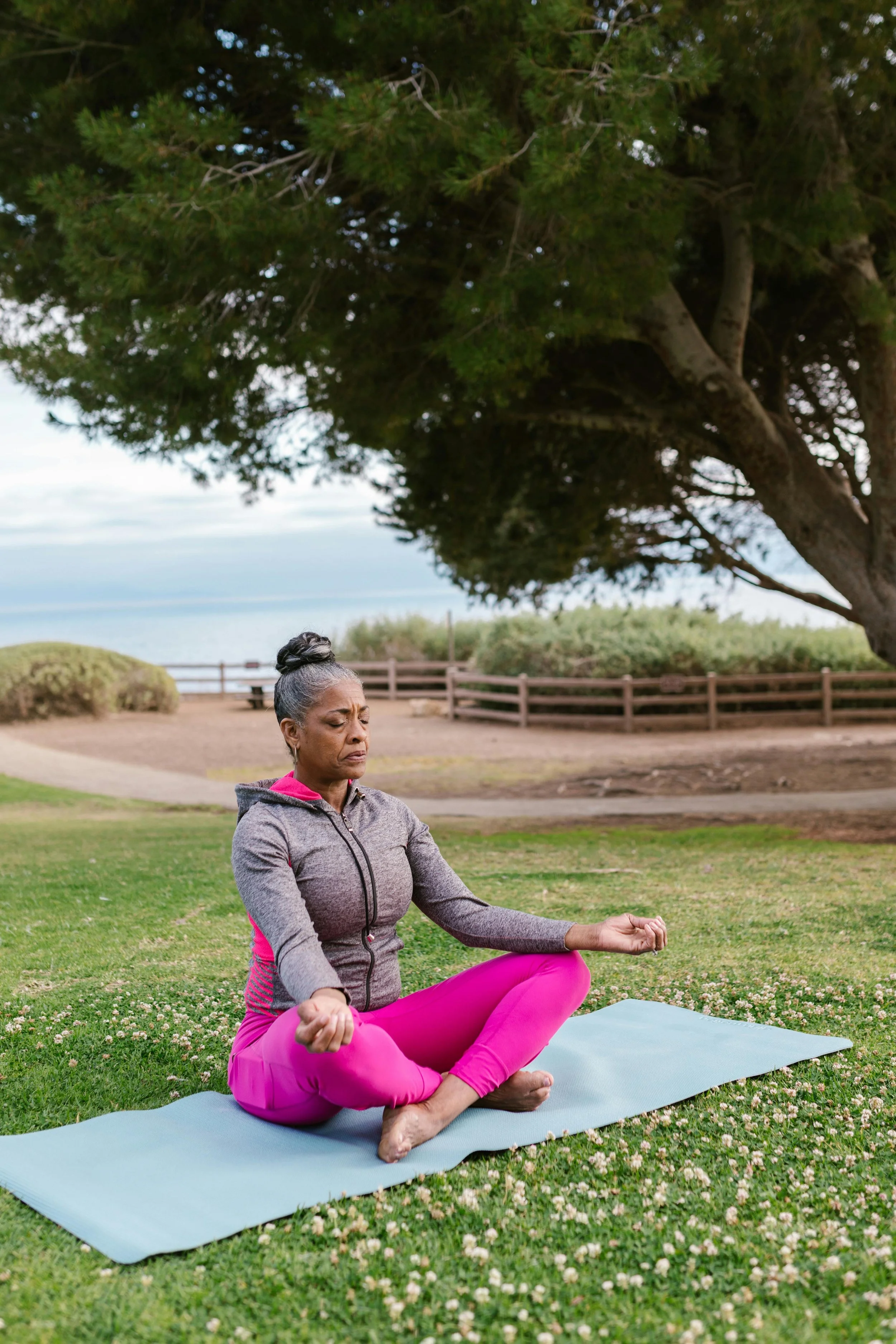 An elderly woman practicing yoga outdoors on a mat under a large tree with green grass and a wooden fence in the background reminding us of the power of hypnosis for anxiety